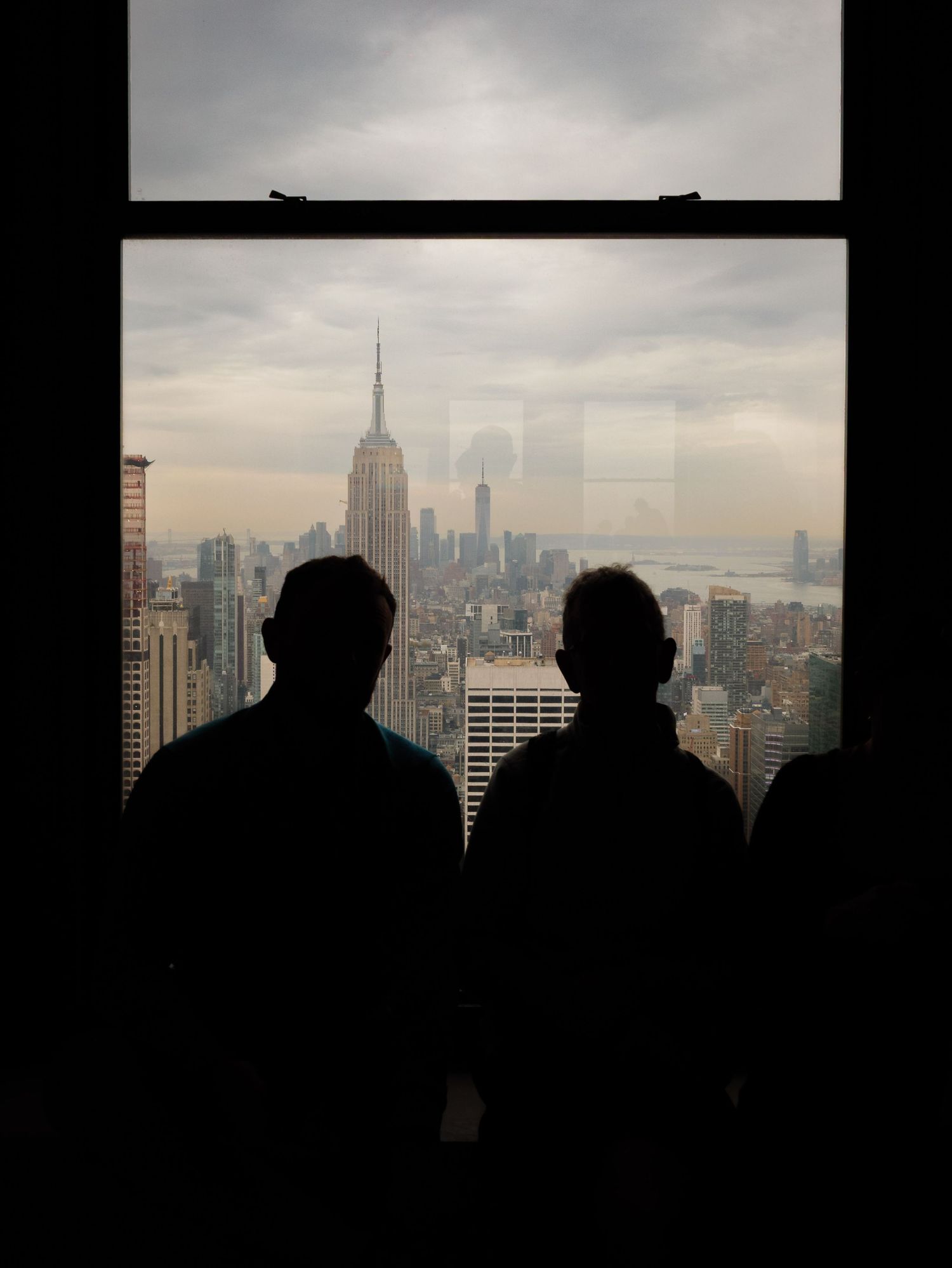 Two silhouetted figures gaze at the New York City skyline featuring the Empire State Building through a large window.