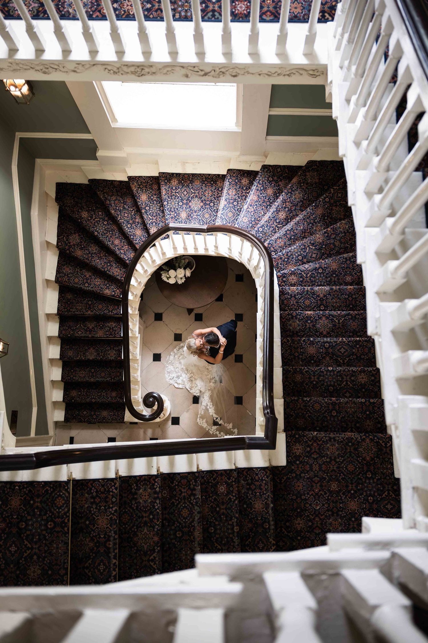 Aerial view of a bride and groom standing at the base of an elegant spiral staircase with dark patterned carpet. Dunboyne Castle