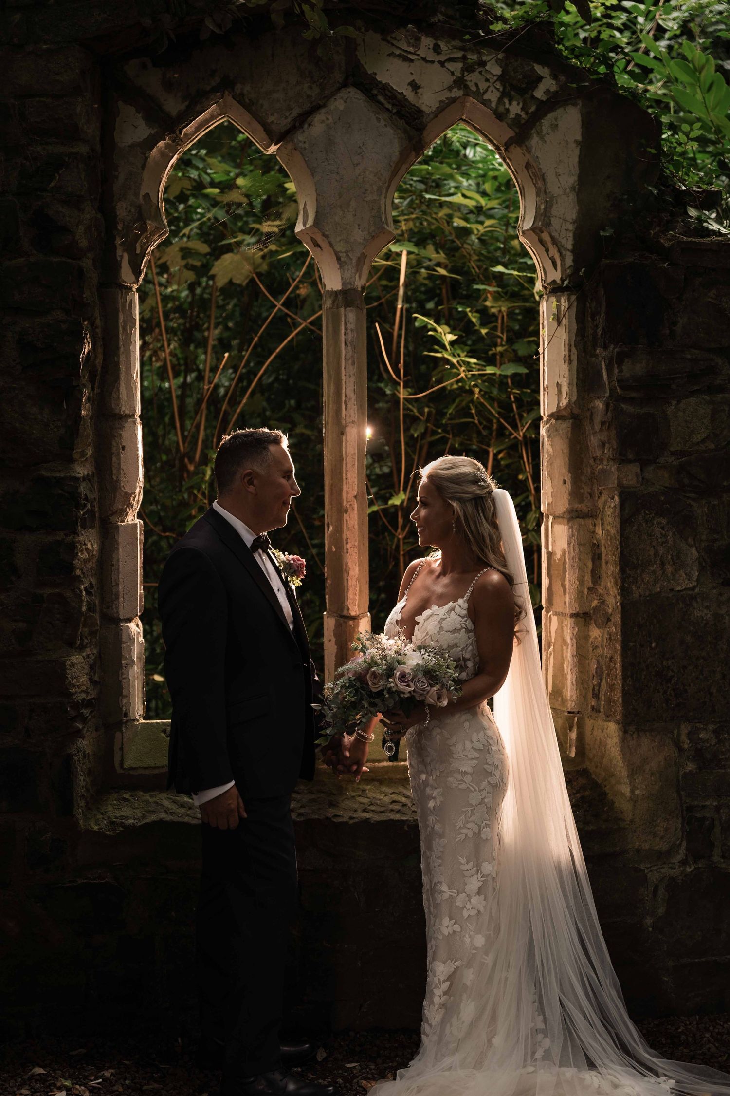 Bride and groom stand before Gothic stone arches surrounded by lush greenery in a romantic, dramatic wedding portrait. Dunboyne Castle
