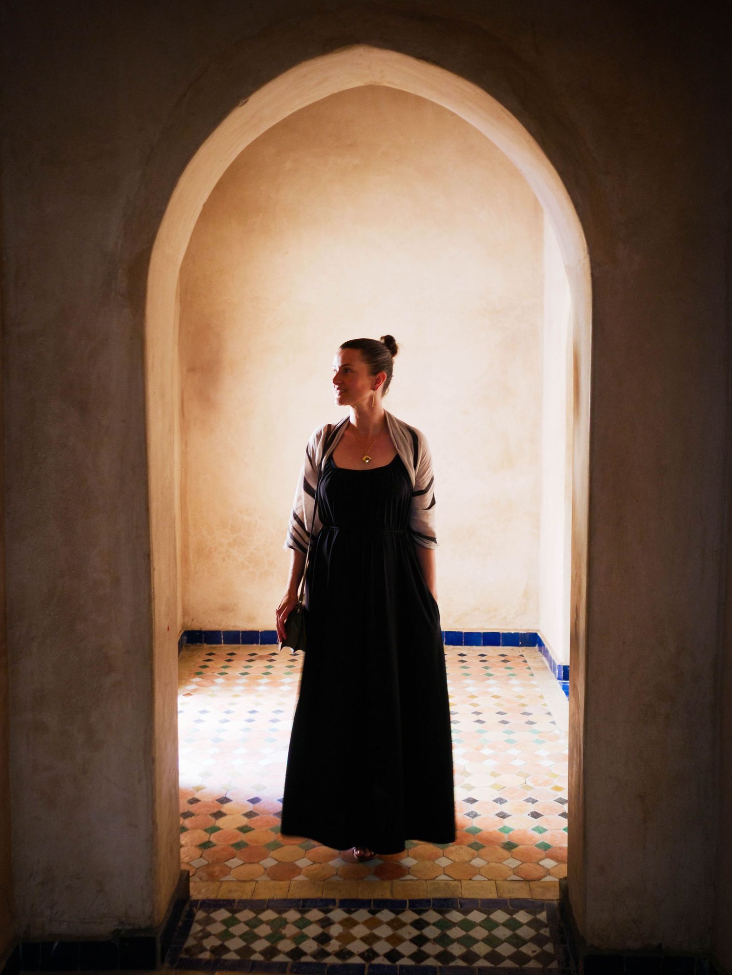 A woman in a black maxi dress stands in a Moorish arched doorway with ornate tiled floors.