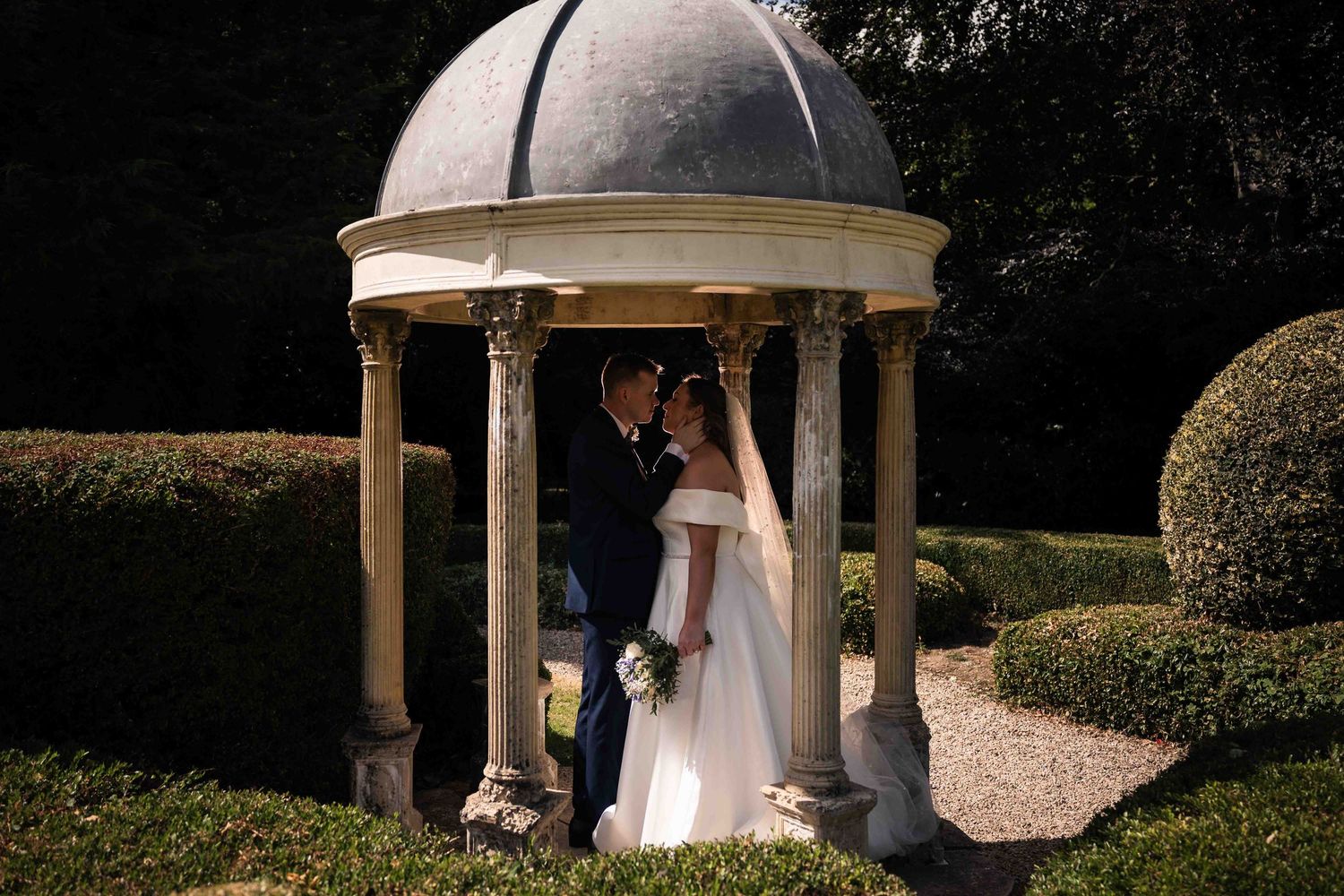 Bride and groom sharing a romantic moment under an elegant stone gazebo surrounded by manicured hedges. Dunboyne Castle