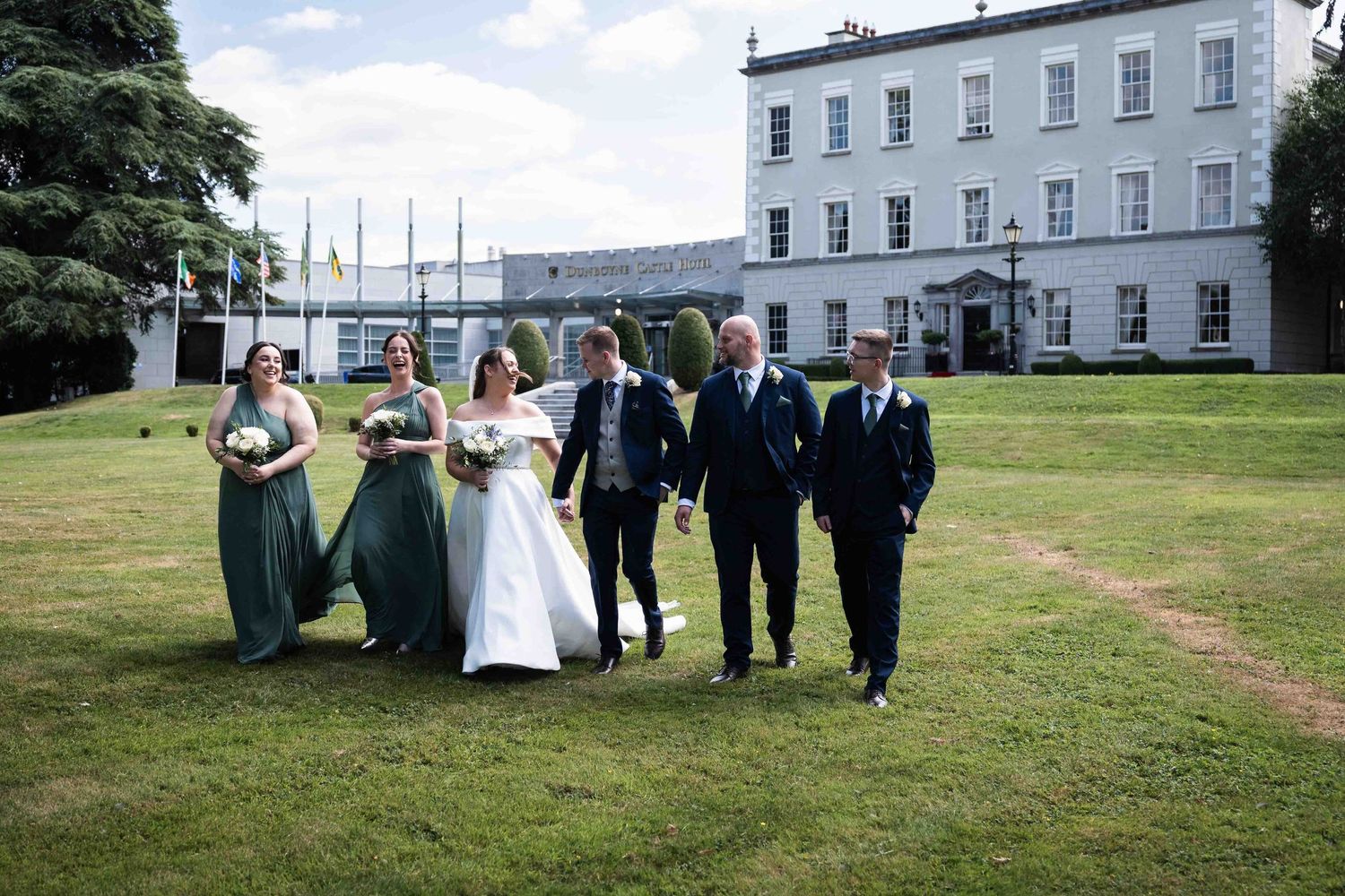 Wedding party at Dunboyne Castle walks on green lawn before grand manor house, bride in white gown with bridesmaids in sage green dresses.