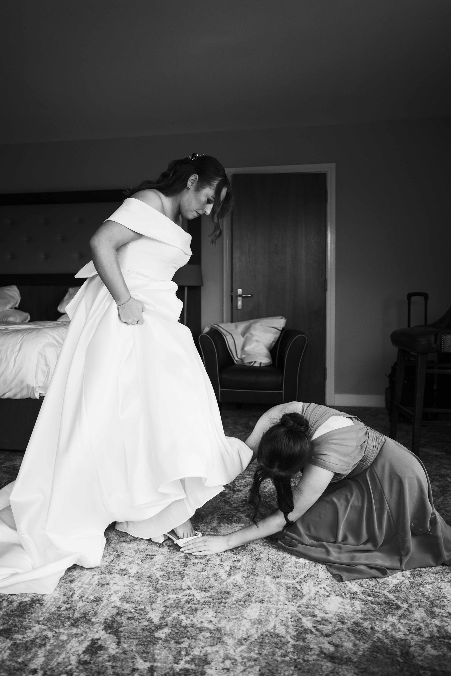 Black and white photo of bridesmaid kneeling to help bride with her shoes in a hotel room. Irish wedding photography. Dunboyne Castle