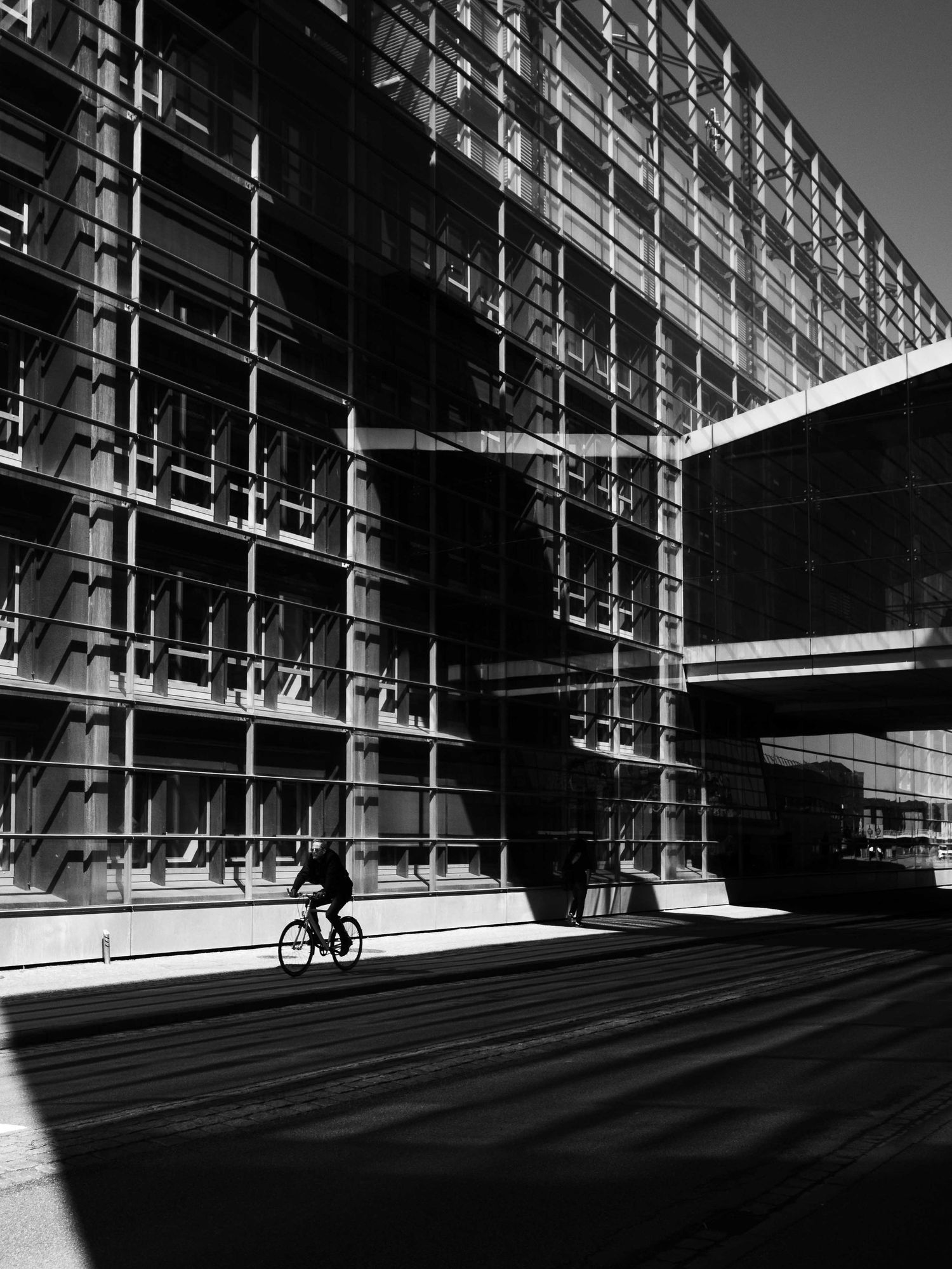 Cyclist silhouette passing modern glass building with dramatic shadows in black and white urban street photography.