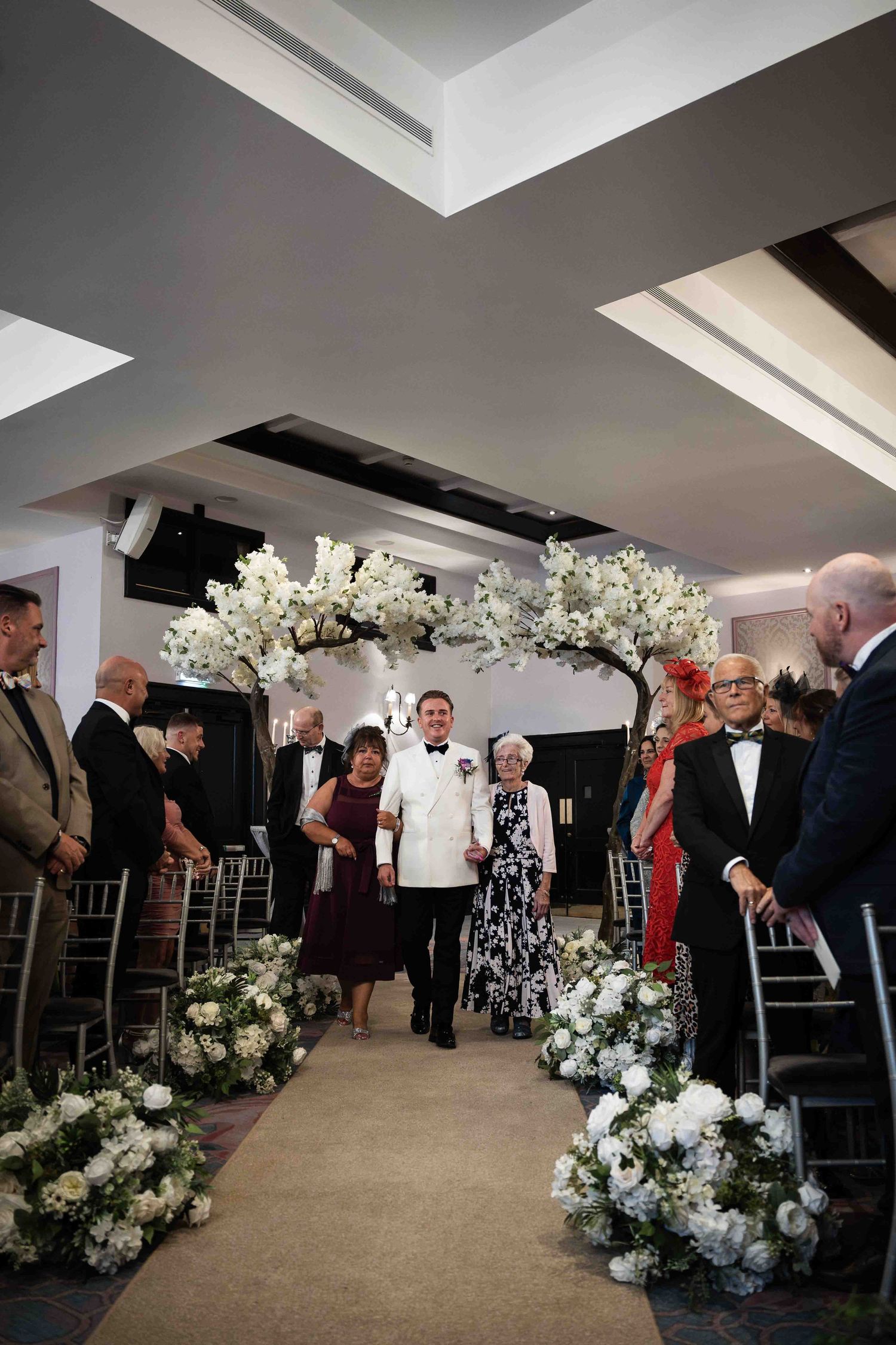 Groom walks down the aisle escorted by family beneath a white cherry blossom floral arch. Queer wedding. Clontarf Castle