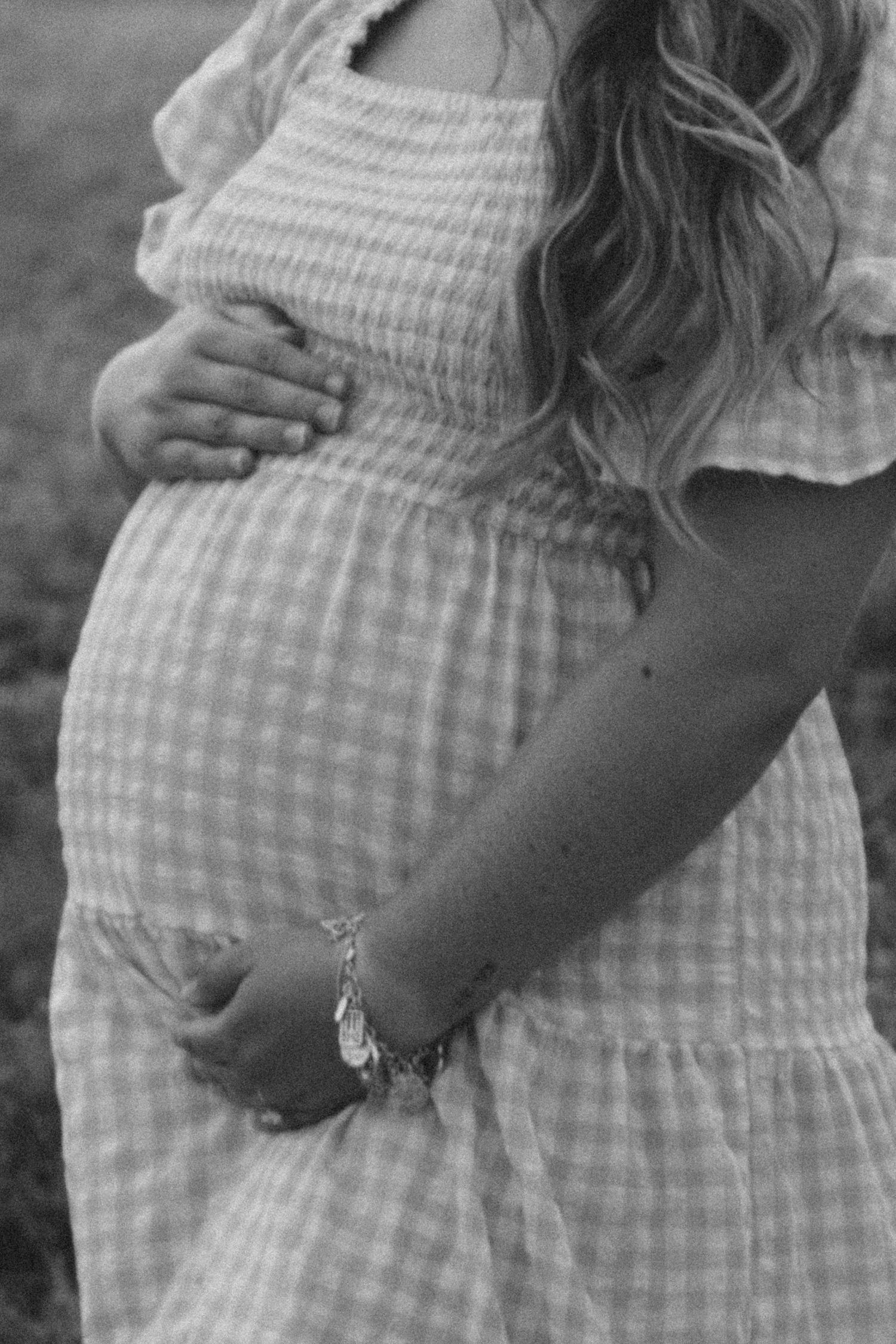 Black and white maternity photo of pregnant woman in gingham dress cradling baby bump with charm bracelet.