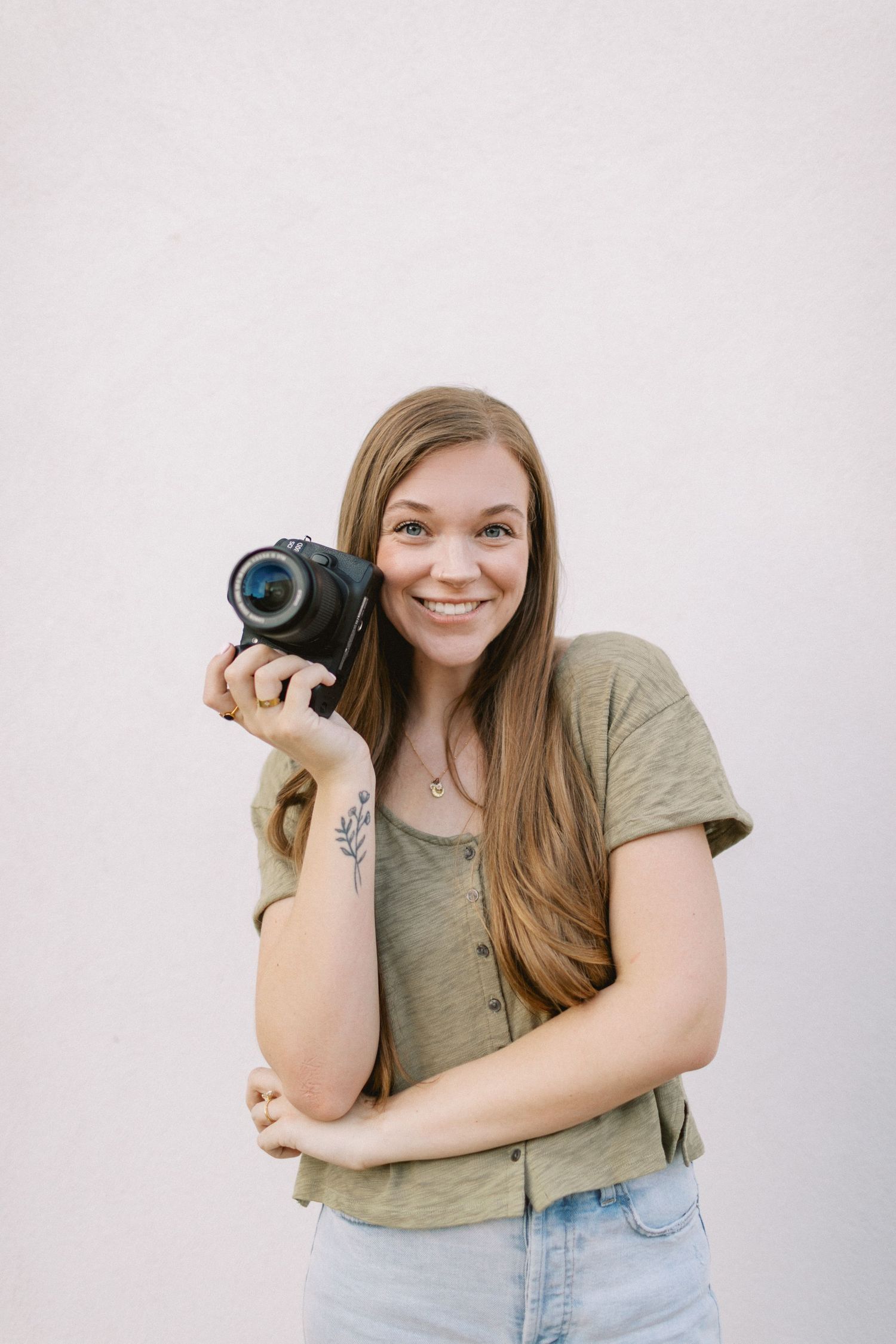 Smiling woman with long brown hair holding a DSLR camera, wearing a green top and jeans against a white wall.