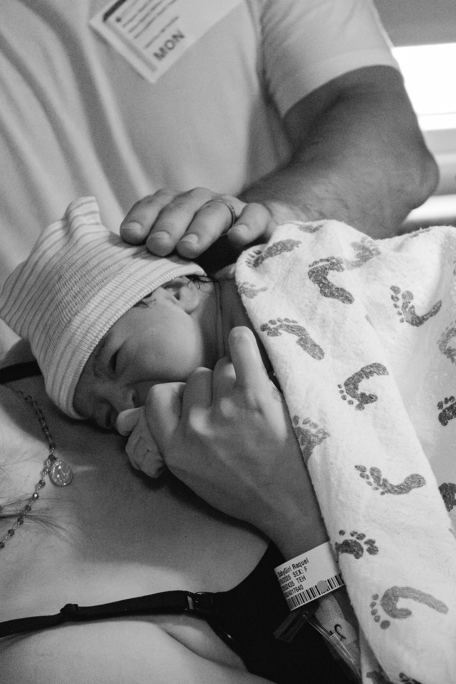 Black and white photo of a newborn baby in a striped hat and footprint blanket being held by an adult in a hospital.