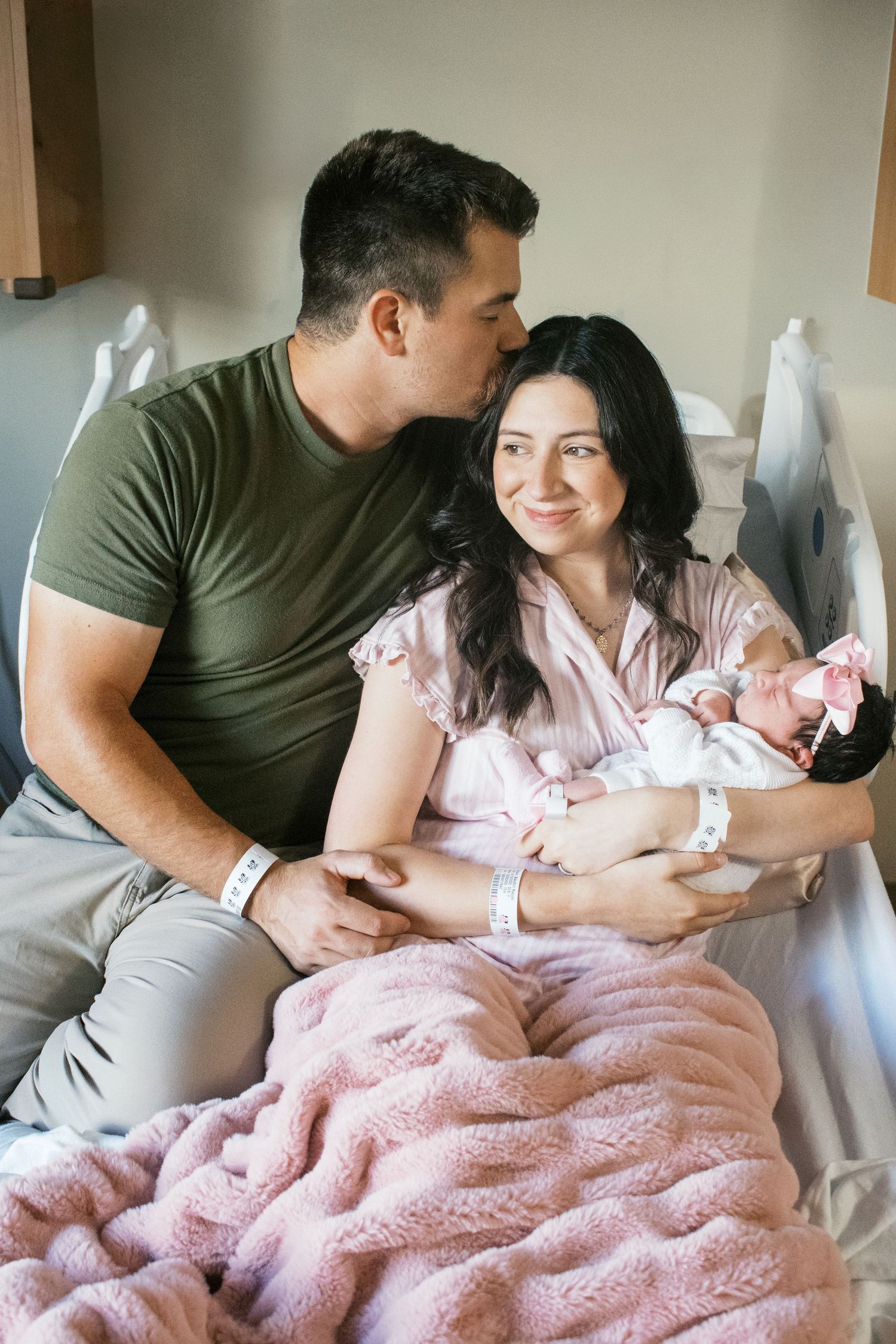 New parents smile in hospital bed, father kissing mother's head as she holds their newborn in pink blanket.