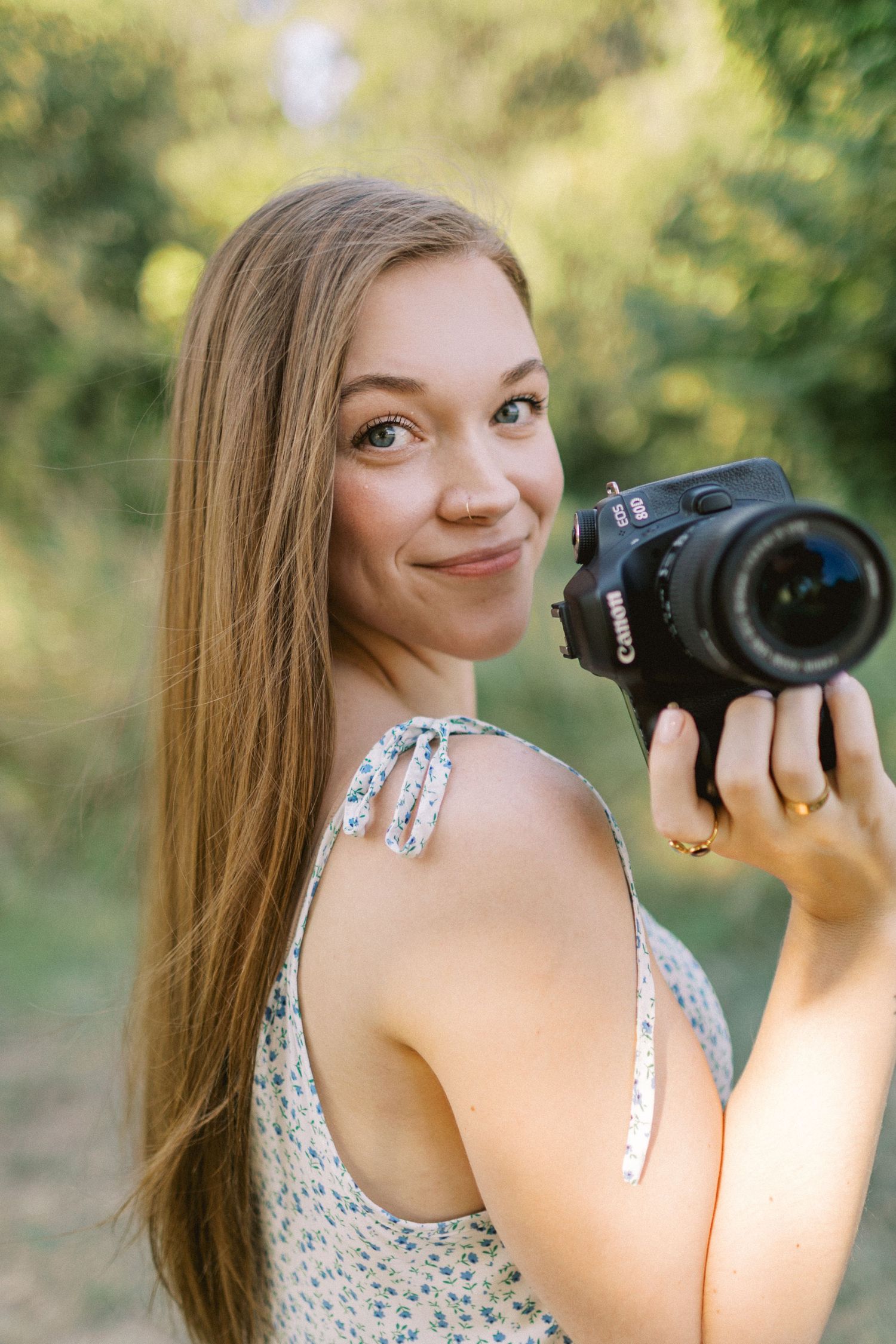 Young woman smiling and holding a Canon DSLR camera outdoors, wearing a floral sundress with green trees in background.