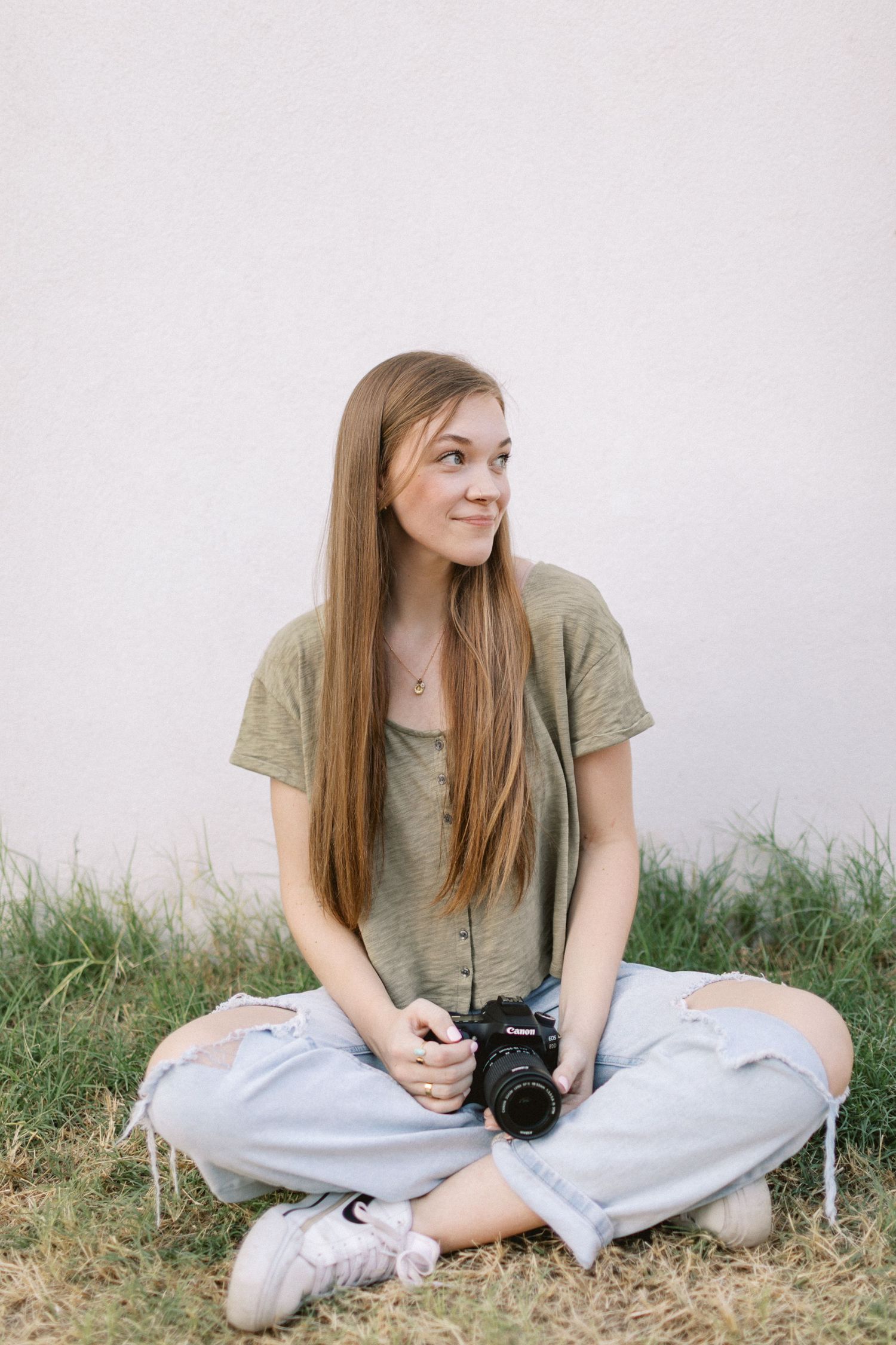 Young woman with long hair sits cross-legged on grass holding a Canon DSLR camera, wearing ripped jeans and olive top.