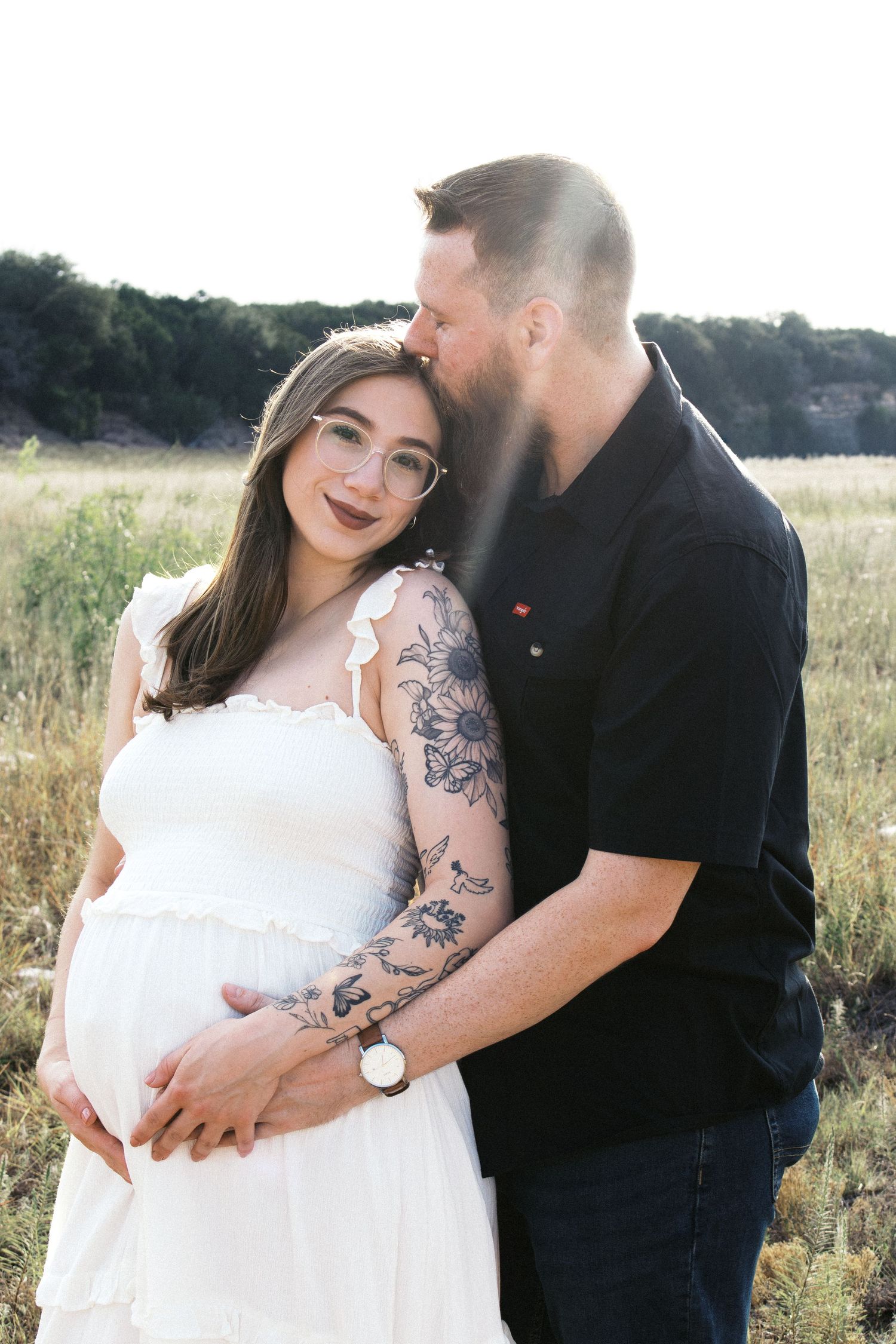 Pregnant woman in white dress with tattooed man in black shirt embracing outdoors in a golden hour maternity photo.