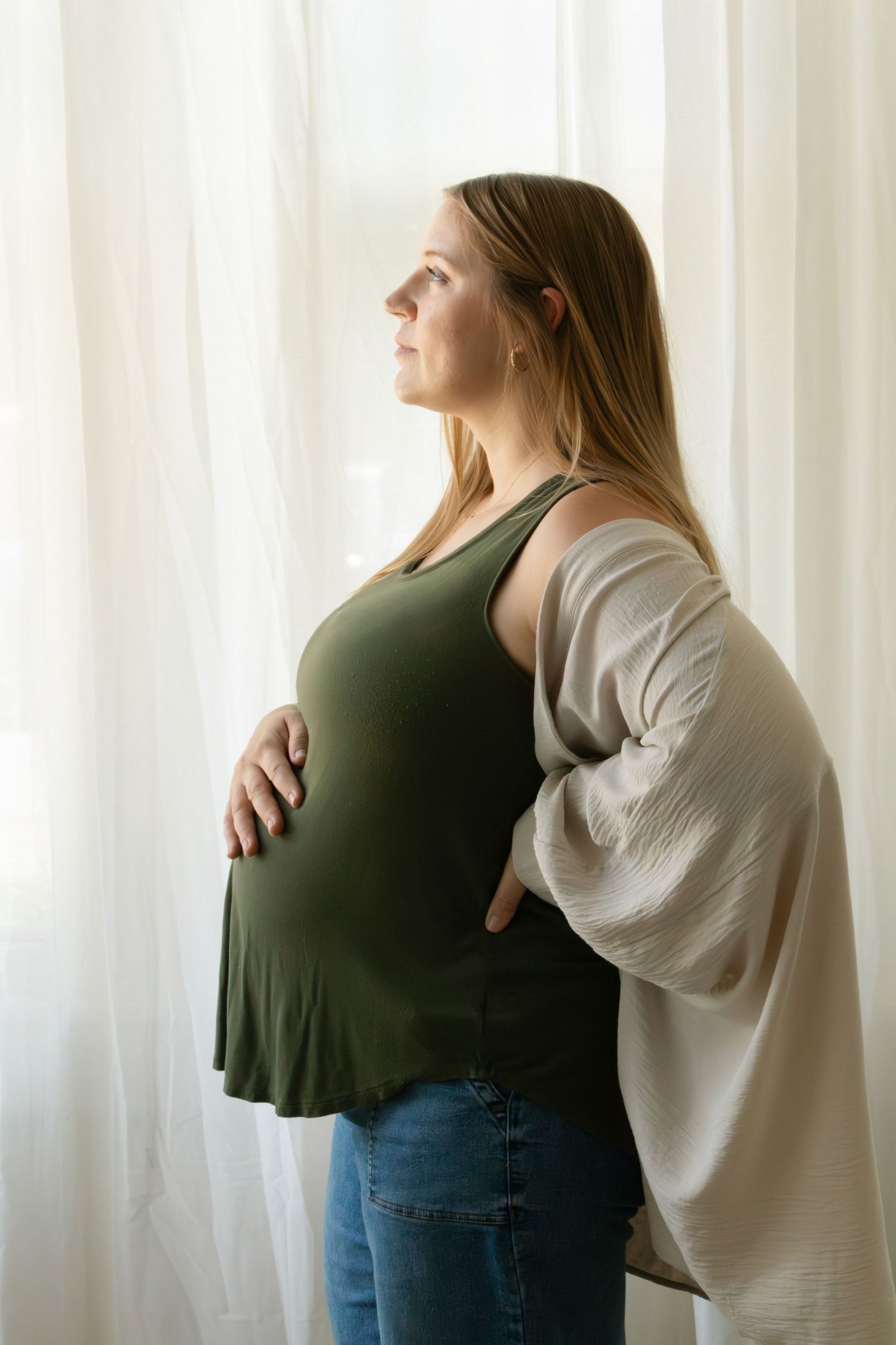 Pregnant woman in green tank top and jeans cradling her baby bump while gazing out a bright window.