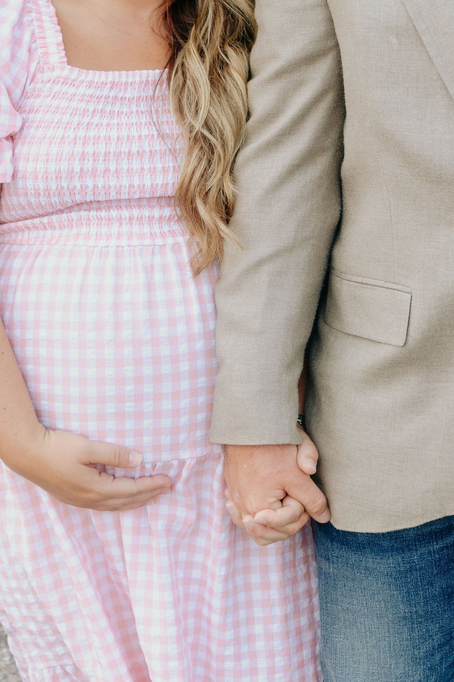 Pregnant woman in pink gingham dress holds hands with partner in tan blazer during maternity photo session.