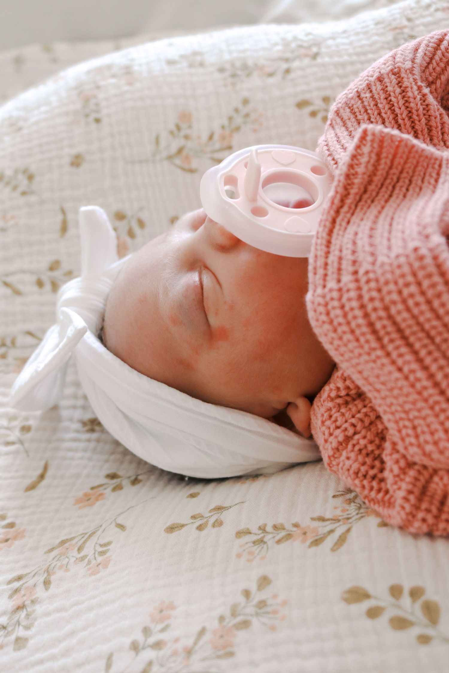 Sleeping newborn baby girl wearing a white bow headband and pink knit hat with a pink pacifier on a floral blanket.