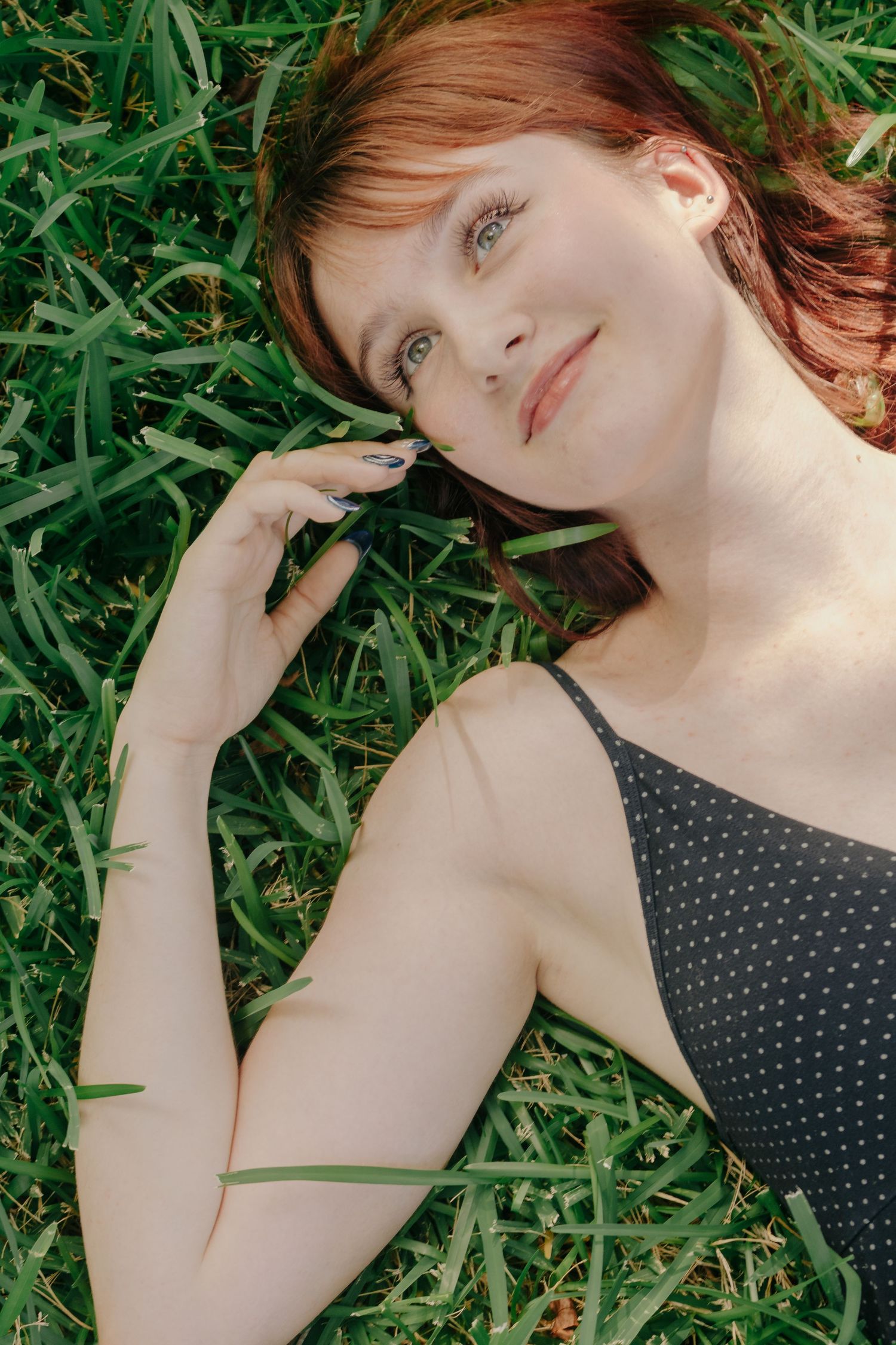 A young redhead woman with blue eyes lying on green grass, wearing a black polka dot top, smiling softly.