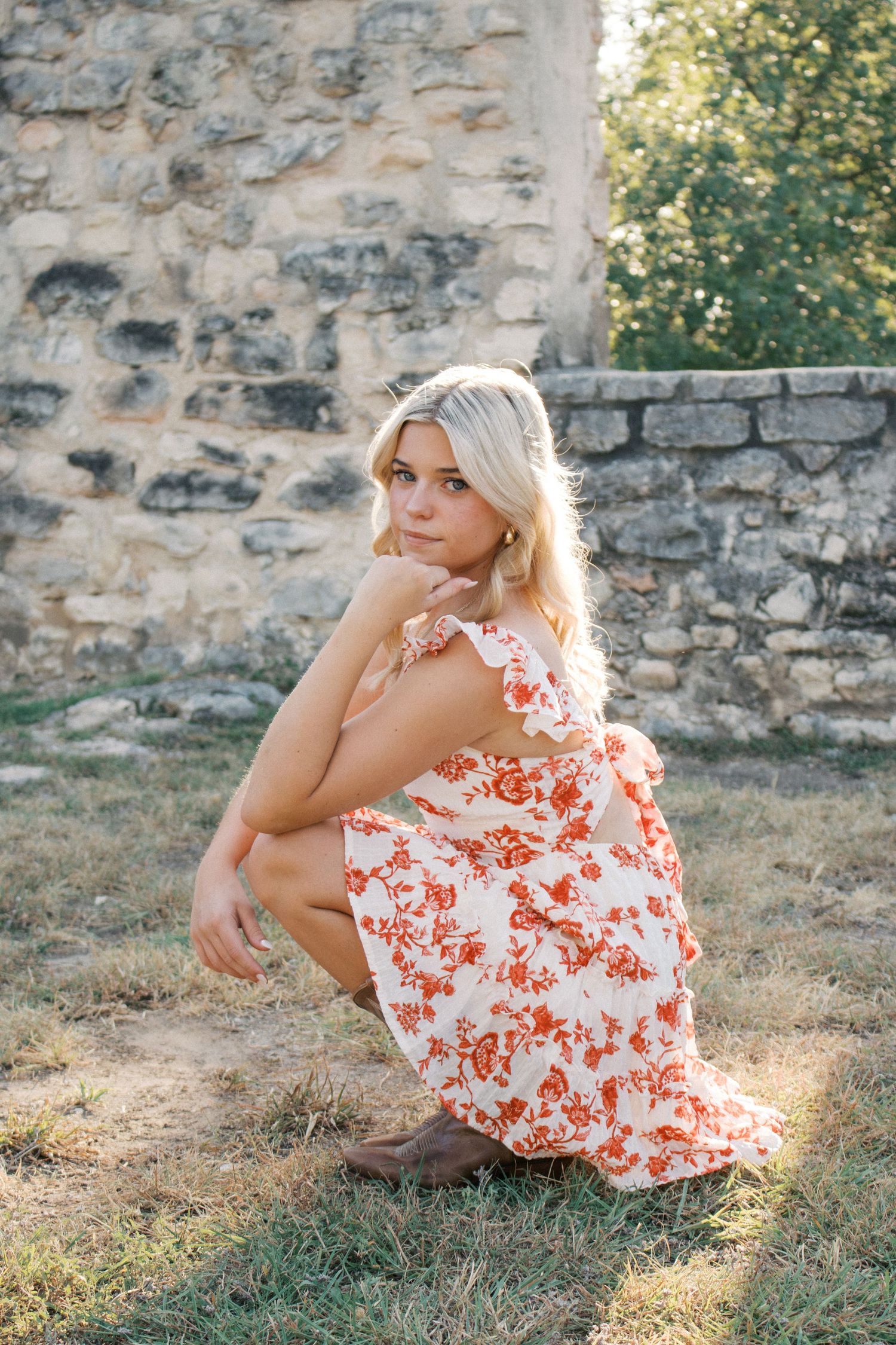 Blonde woman in floral red and white dress crouching on grass near rustic stone wall in golden sunlight.