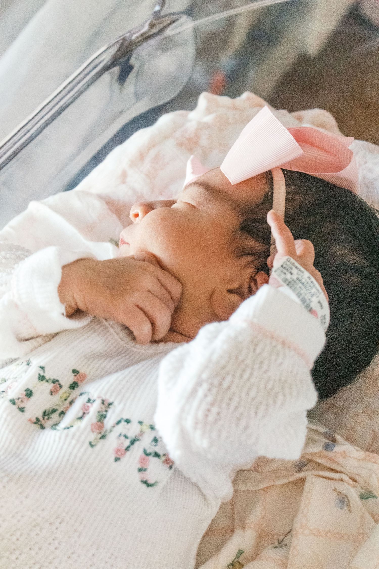 Newborn baby girl sleeping in hospital bassinet wearing white outfit and pink bow headband.