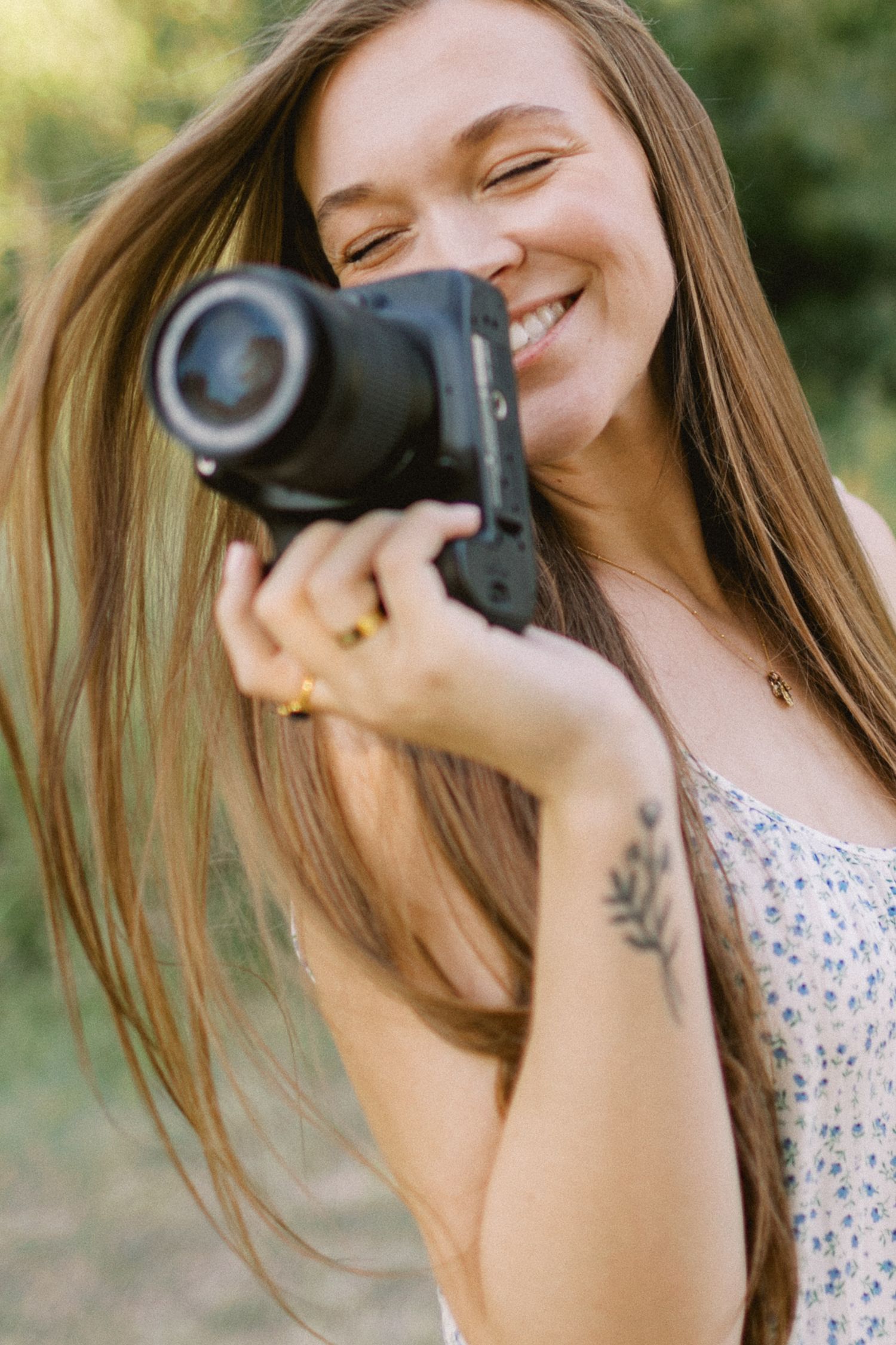 Smiling woman with long hair holding a DSLR camera outdoors, wearing a floral top with a floral arm tattoo.