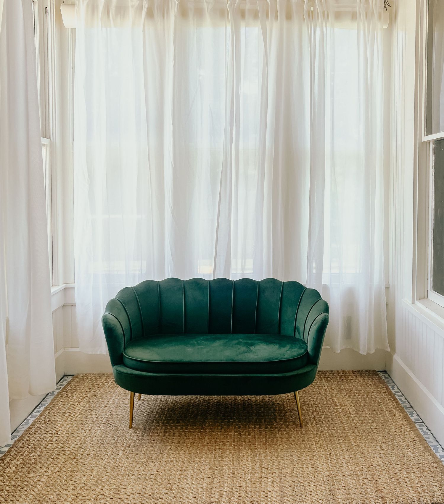 Elegant green velvet shell-shaped loveseat with gold legs on jute rug before sheer white curtains.