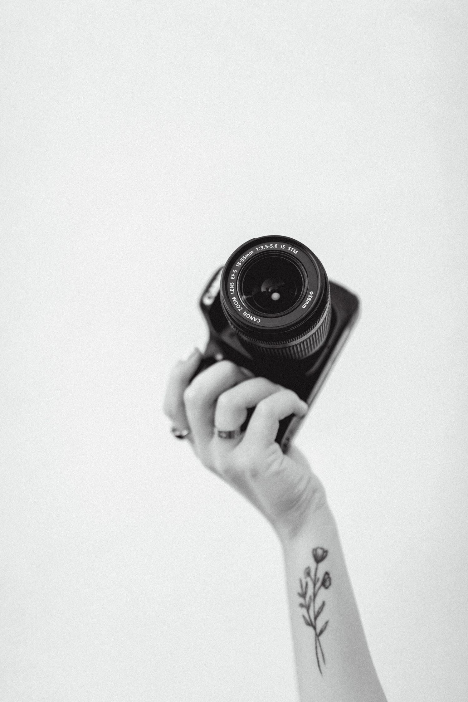 A hand with a floral wrist tattoo holds a DSLR camera up against a white background in black and white.