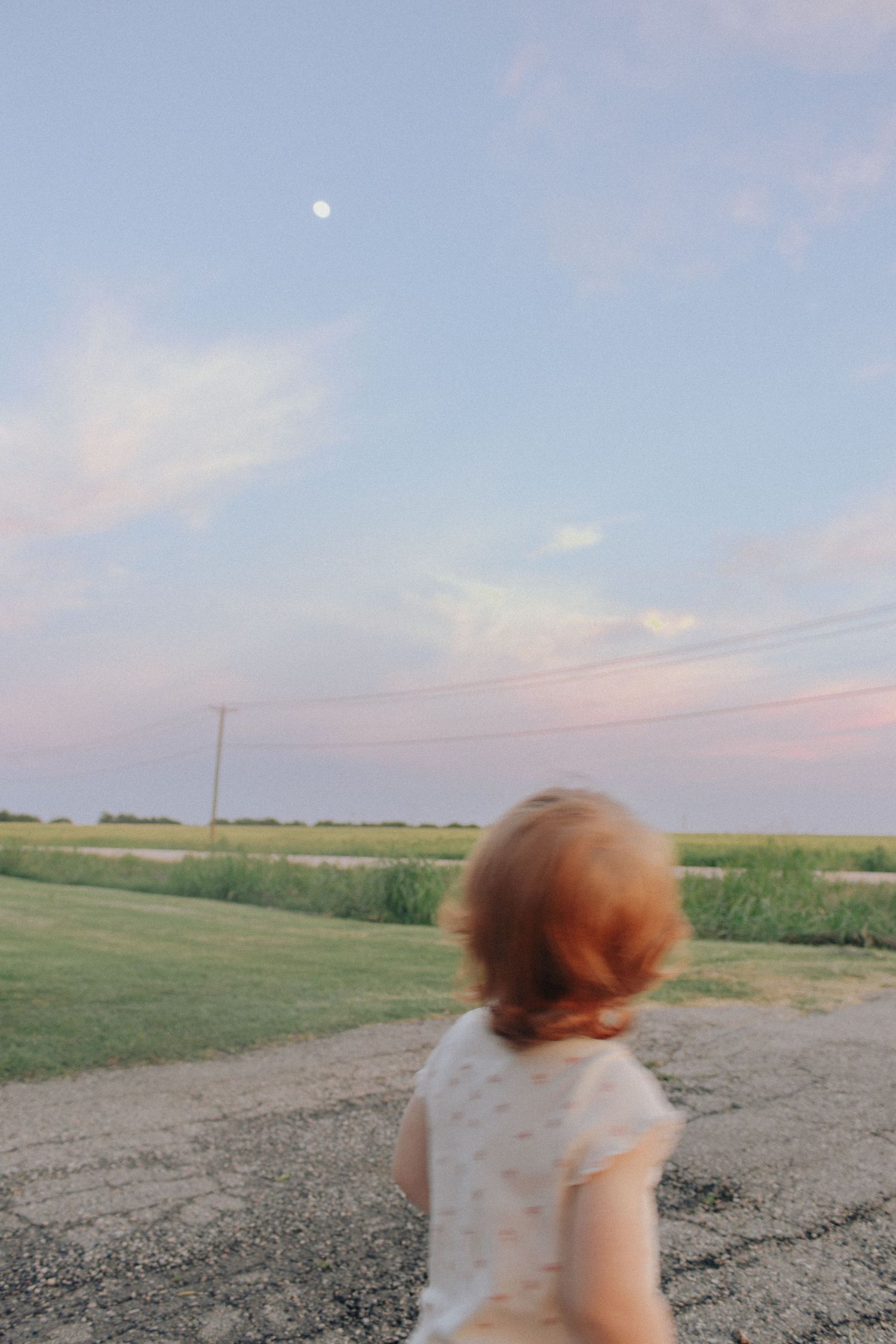 A young red-haired child in a white dress stands on a rural road at dusk, gazing at the moon in a pastel sky.