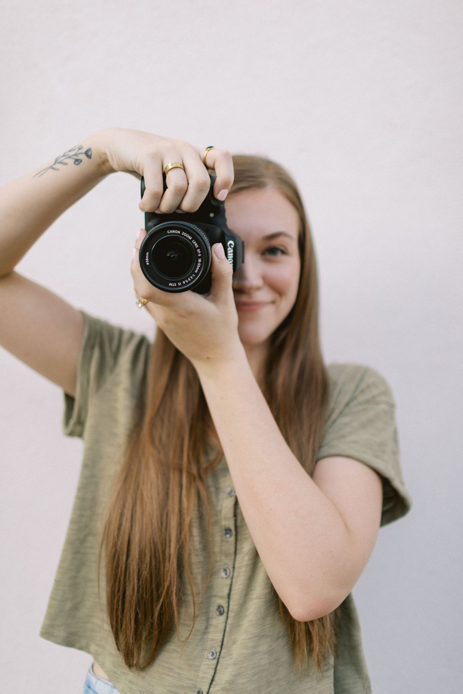 Young woman with long hair smiling while holding a Canon DSLR camera up to her eye outdoors.