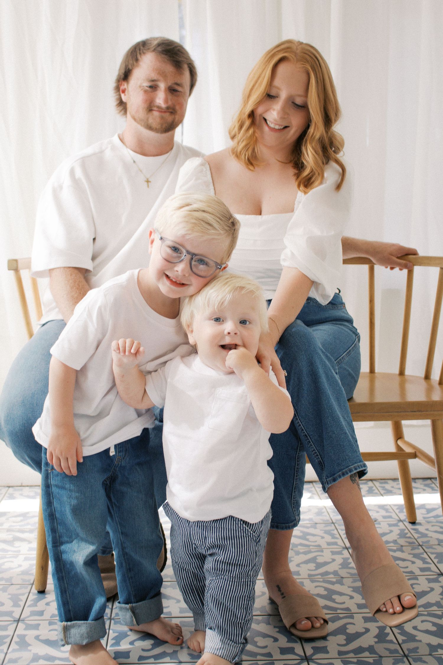 A happy family of four in white tops and jeans poses for a portrait, with two young boys smiling in front of their parents.