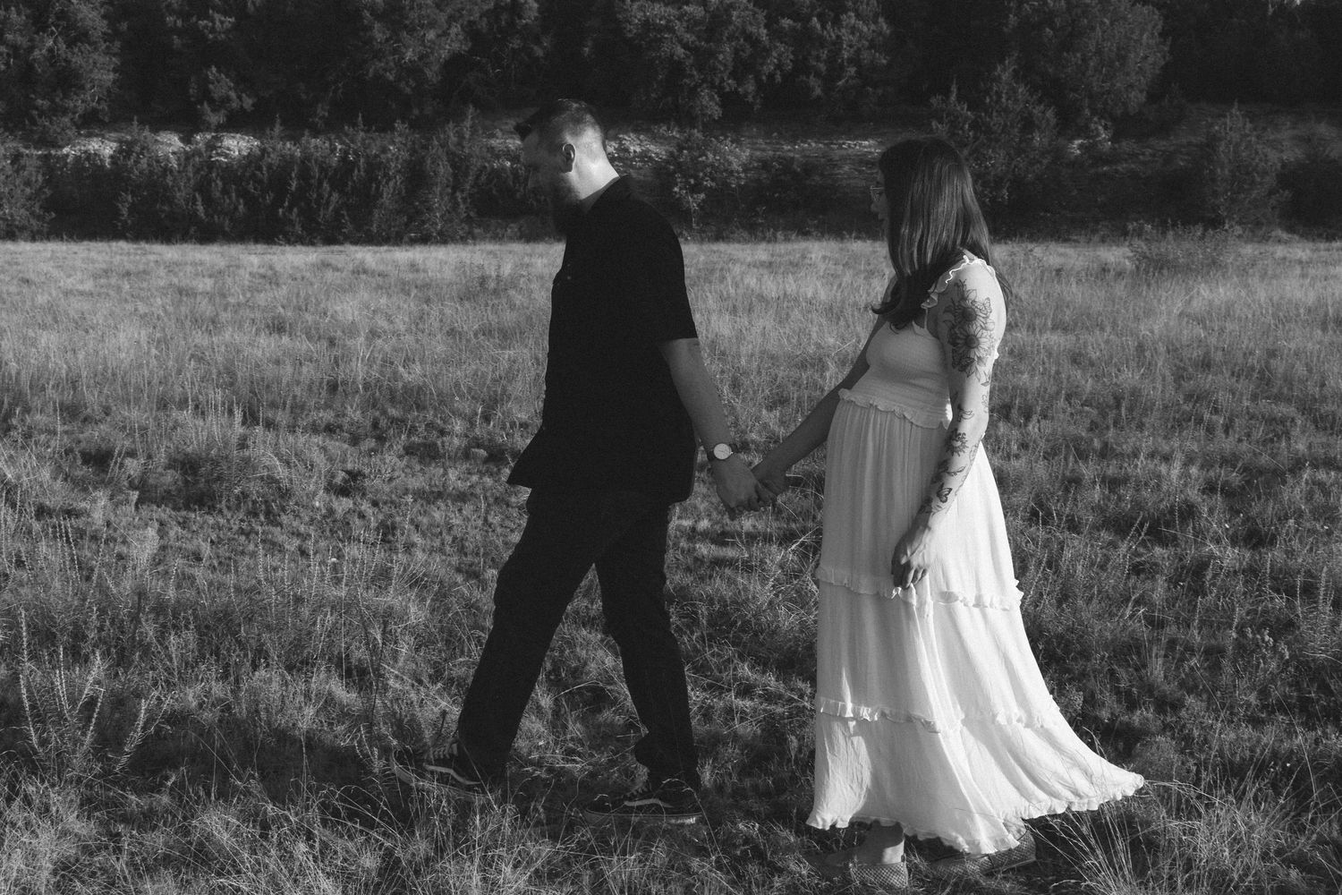 Black and white photo of a couple holding hands walking through a grassy field, woman in flowing white dress.