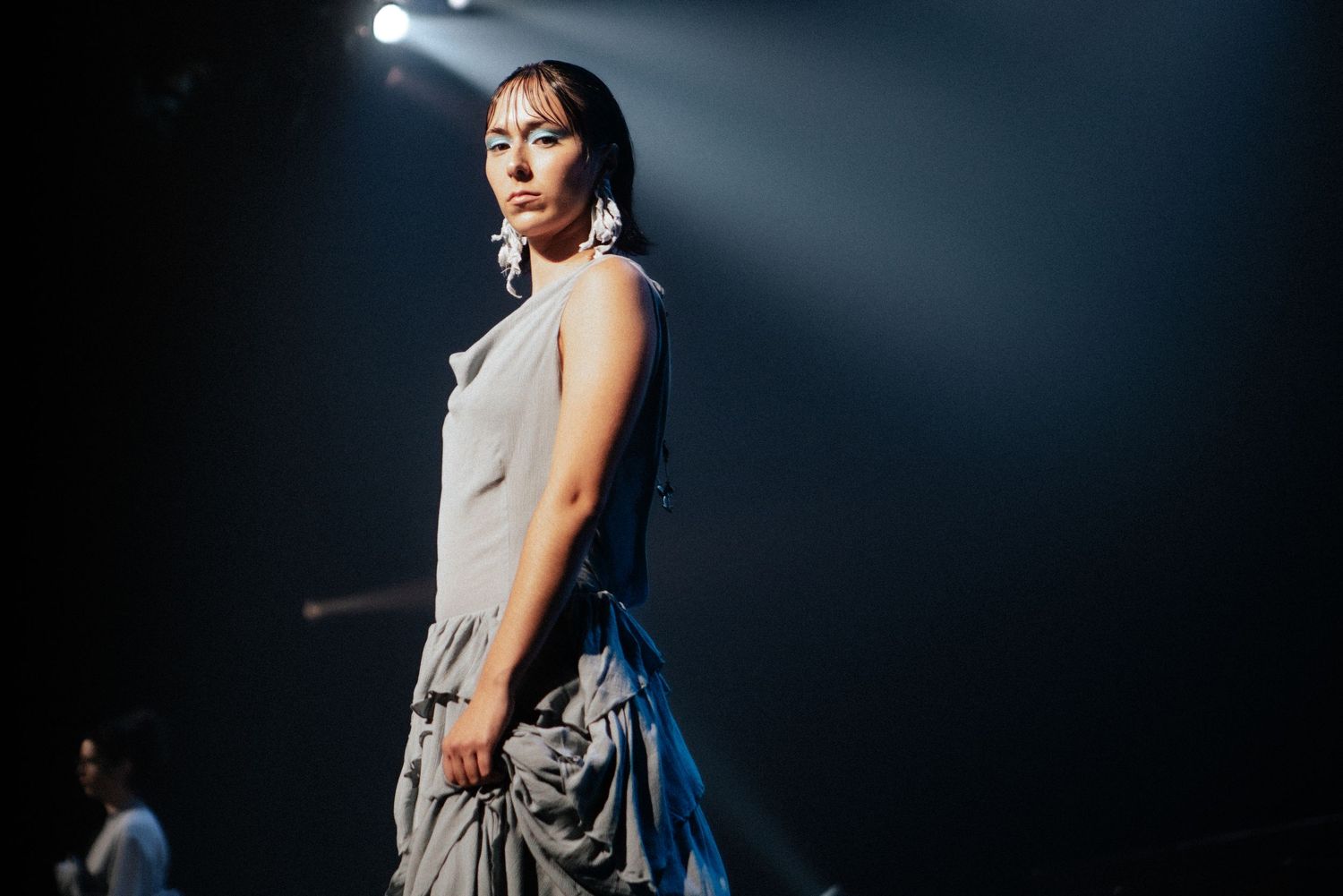 Model in a flowing gray gown walking the runway under moody stage lighting, fashion photography by HAMIDI Photography.