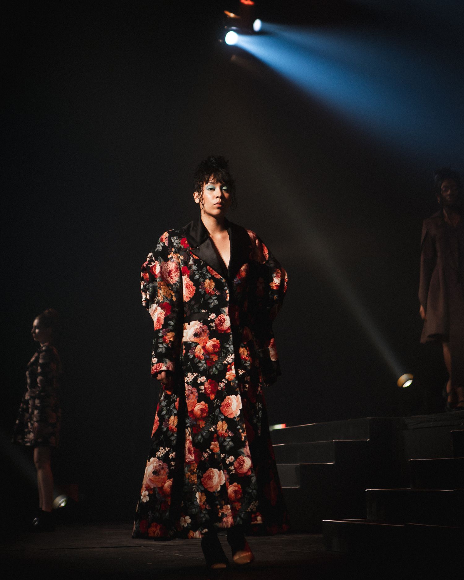 Model on stage in a colorful floral-patterned outfit under dramatic runway lighting, fashion photography by HAMIDI Photography.