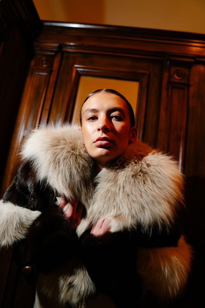 Model in a cream fur-trimmed coat under warm indoor lighting, fashion photography by HAMIDI Photography.