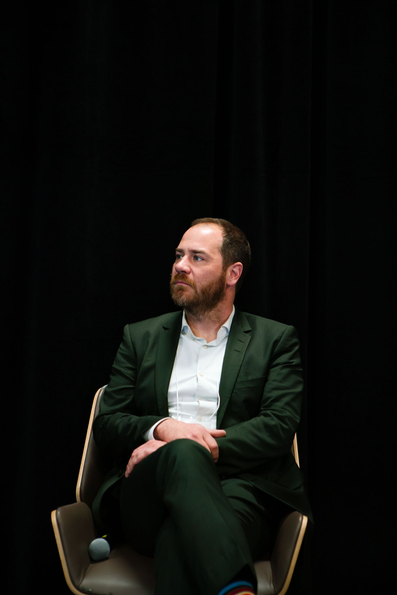 Panelist in a green jacket seated on a dark stage during a conference session, event photography by HAMIDI Photography.