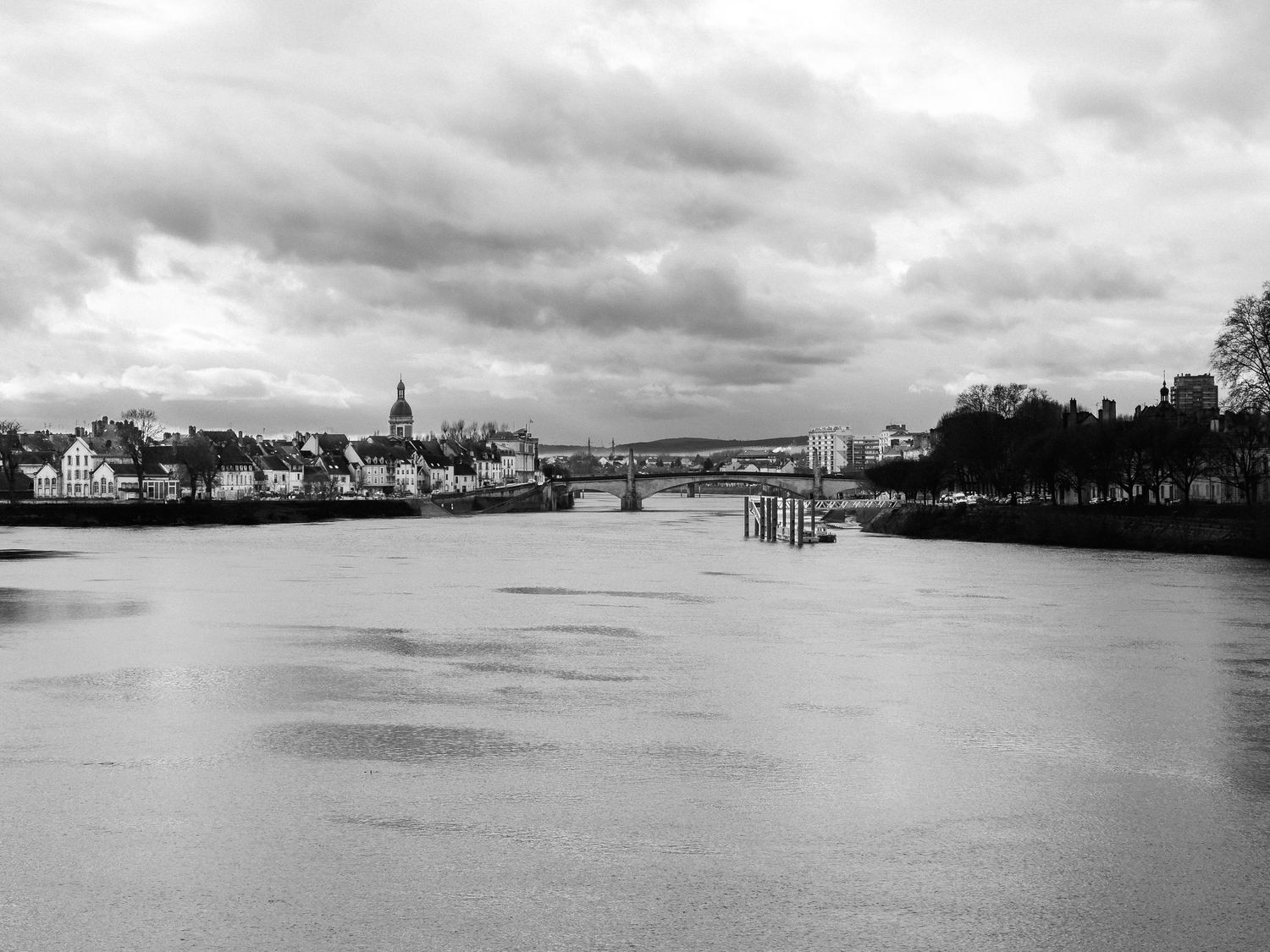 Black and white photo of a river with a bridge, church spire, and cloudy sky in a European town.