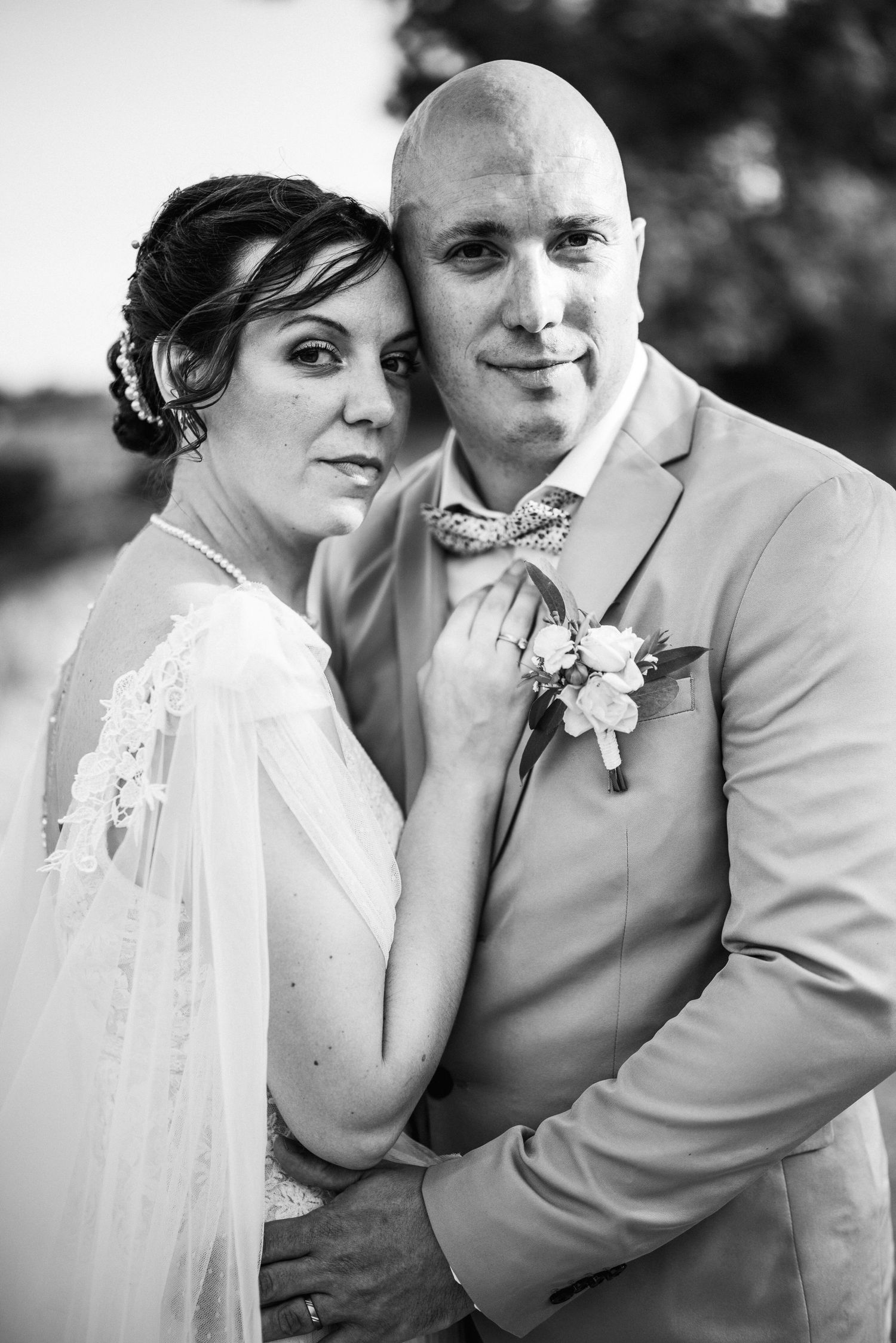 Black and white wedding portrait of a bride in white lace and groom in suit with boutonniere embracing outdoors.