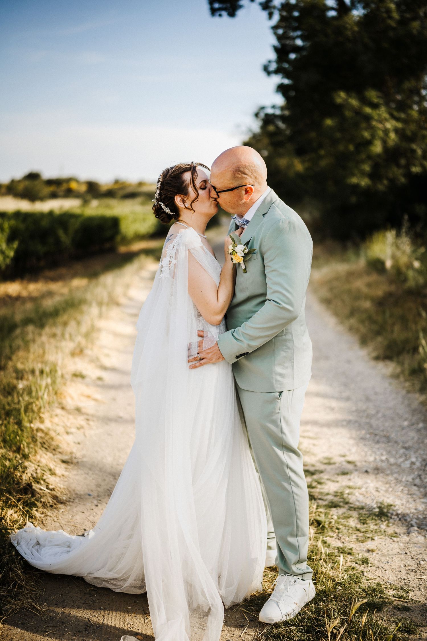 Bride and groom share a kiss on a sunny dirt path, she in a white gown, he in a sage suit with boutonniere.