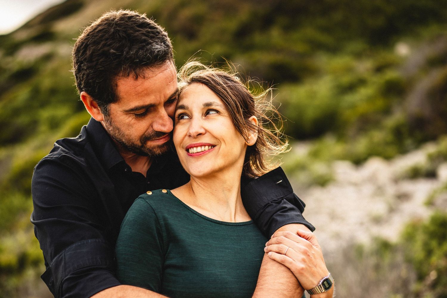 A couple embraces outdoors, the man nuzzling the smiling woman in a green top against a lush hillside.