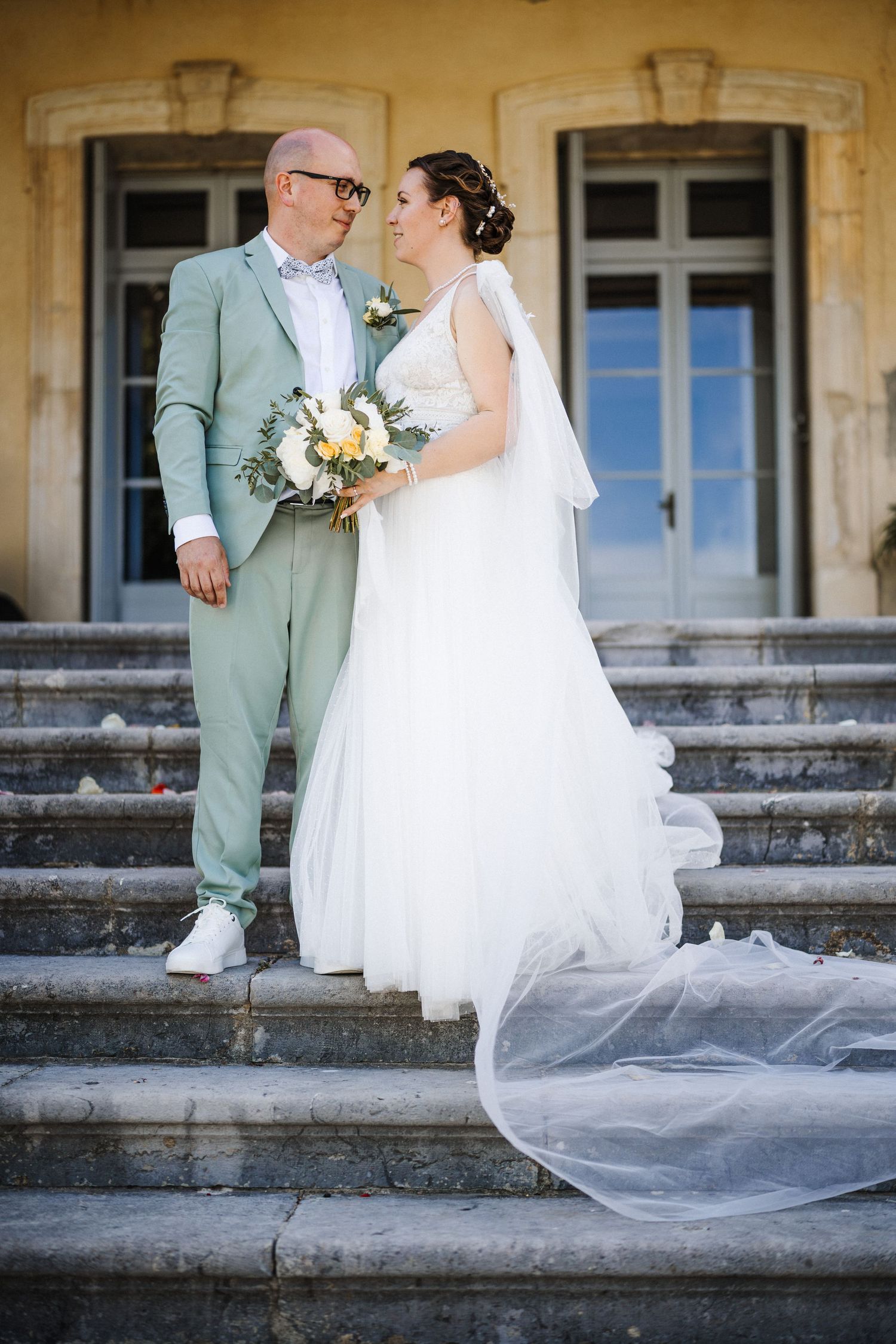 Bride and groom share a romantic moment on stone steps, bride in white gown with veil, groom in mint suit.