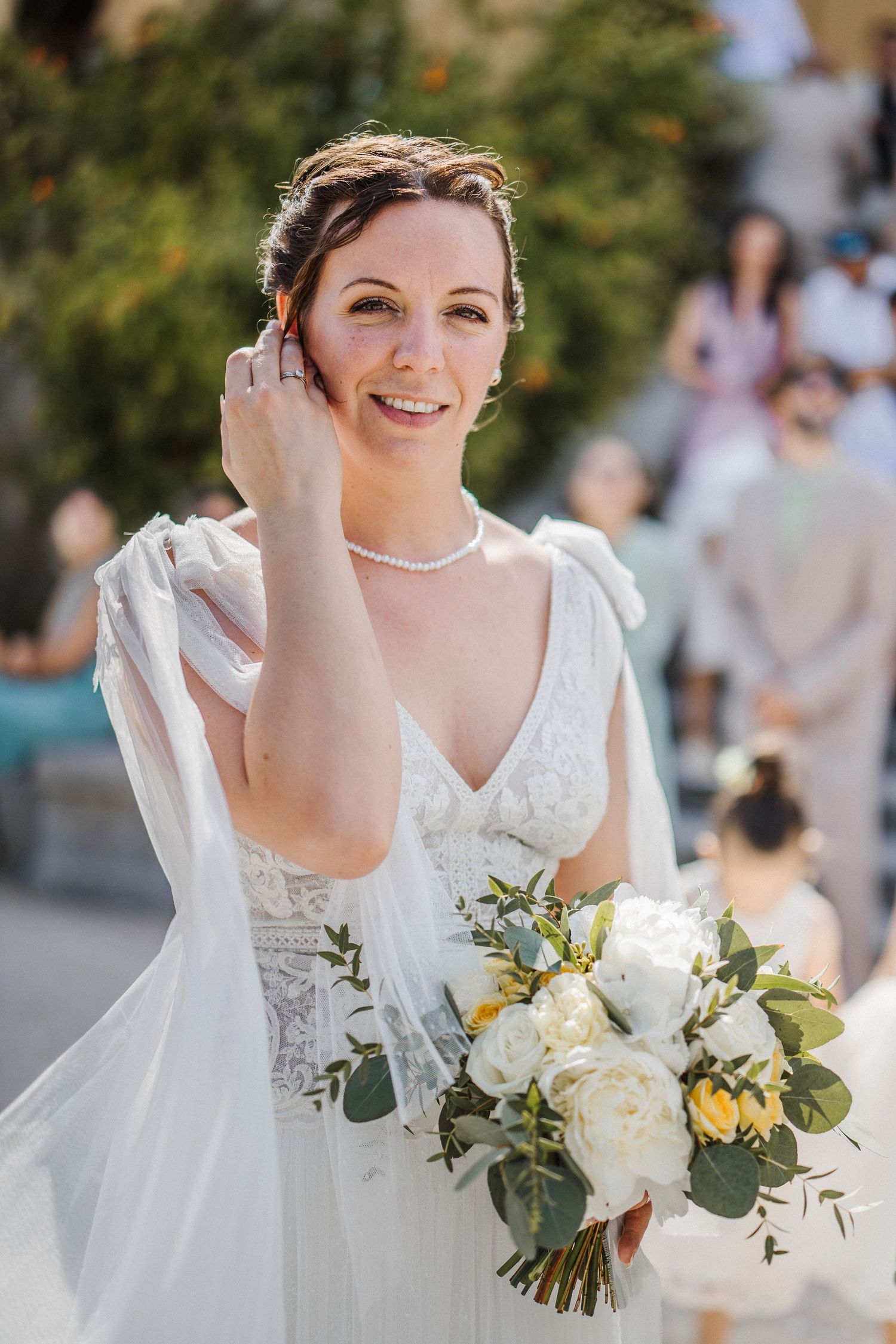 Smiling bride in white lace gown holds bouquet of white roses and greenery, wearing pearl necklace outdoors.