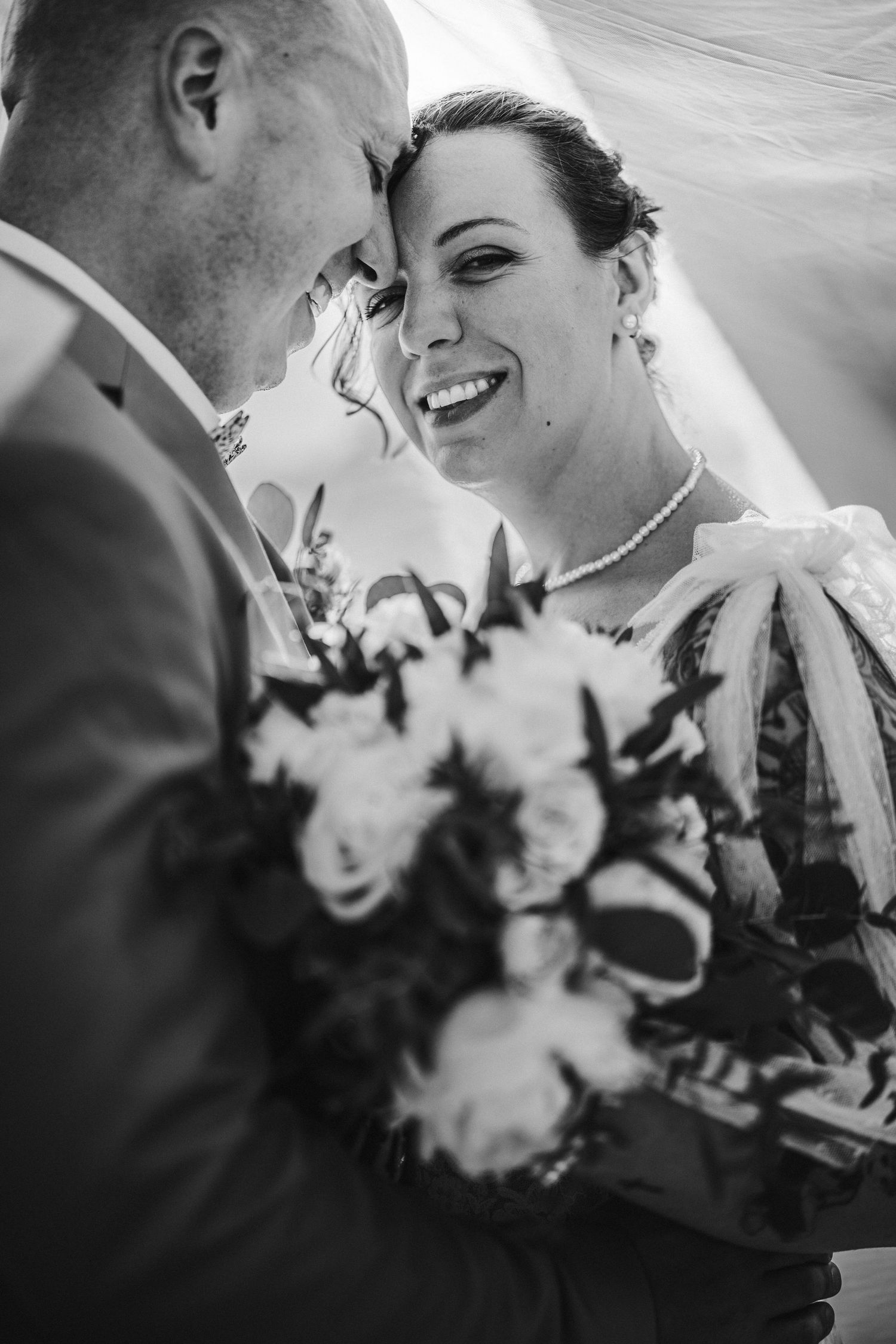 Black and white wedding photo of smiling bride holding bouquet, forehead touching groom in tender romantic moment.