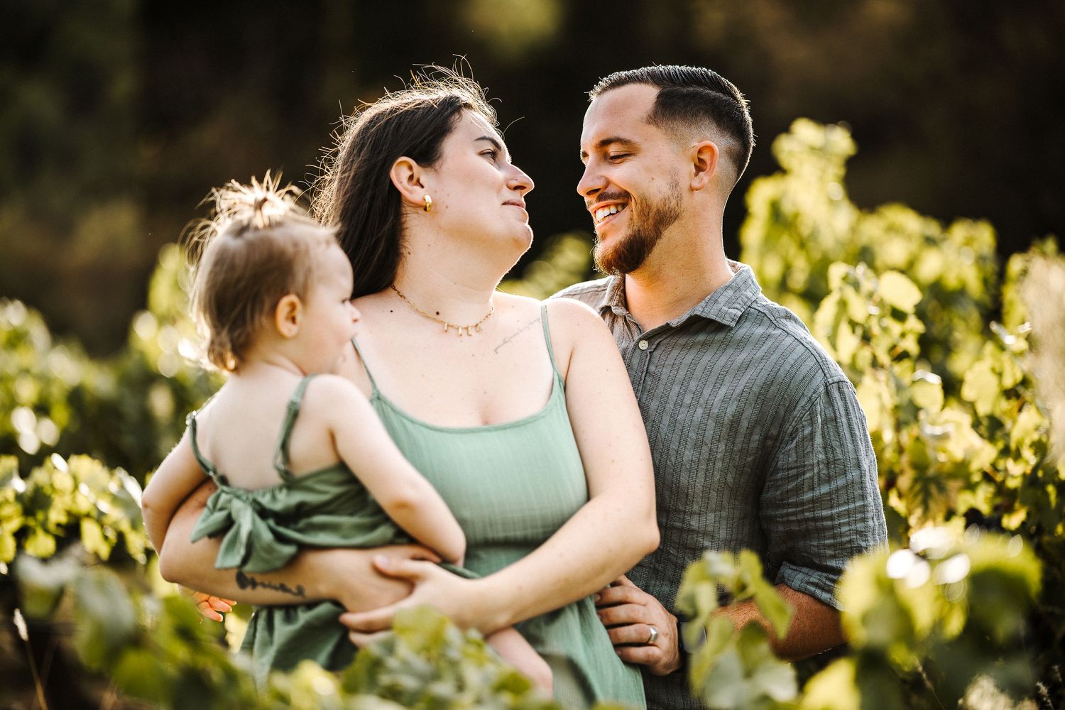 A happy young couple smiles at each other outdoors while holding their baby girl in a lush green vineyard.