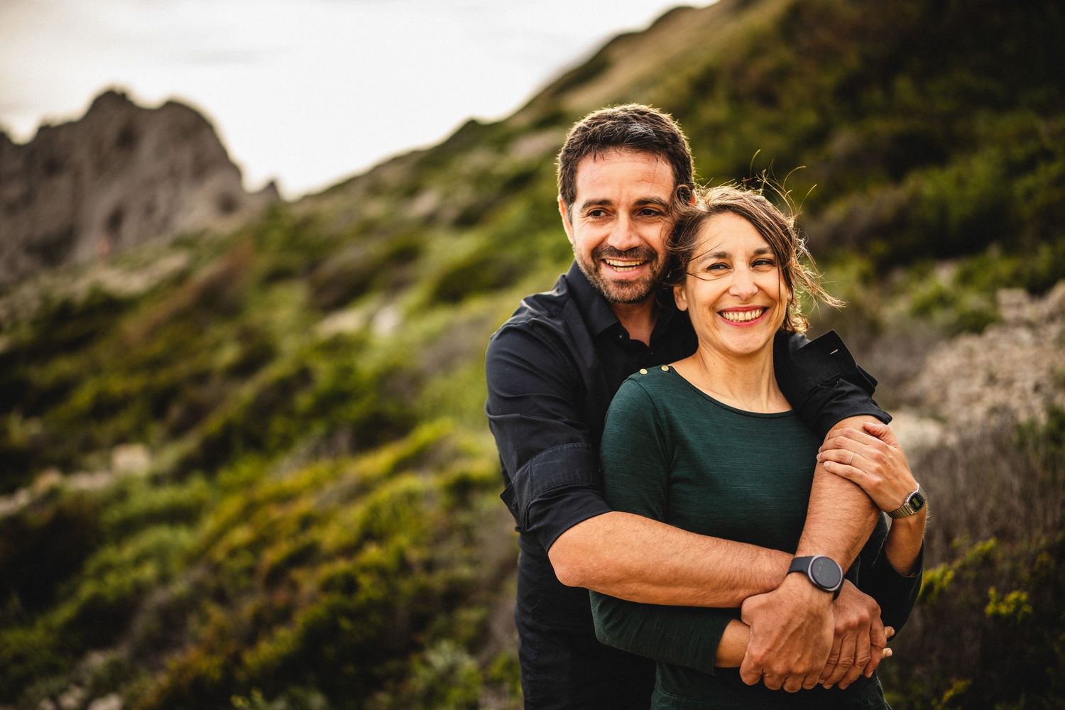 A happy couple hugs closely outdoors, smiling warmly with a scenic green hillside in the background.
