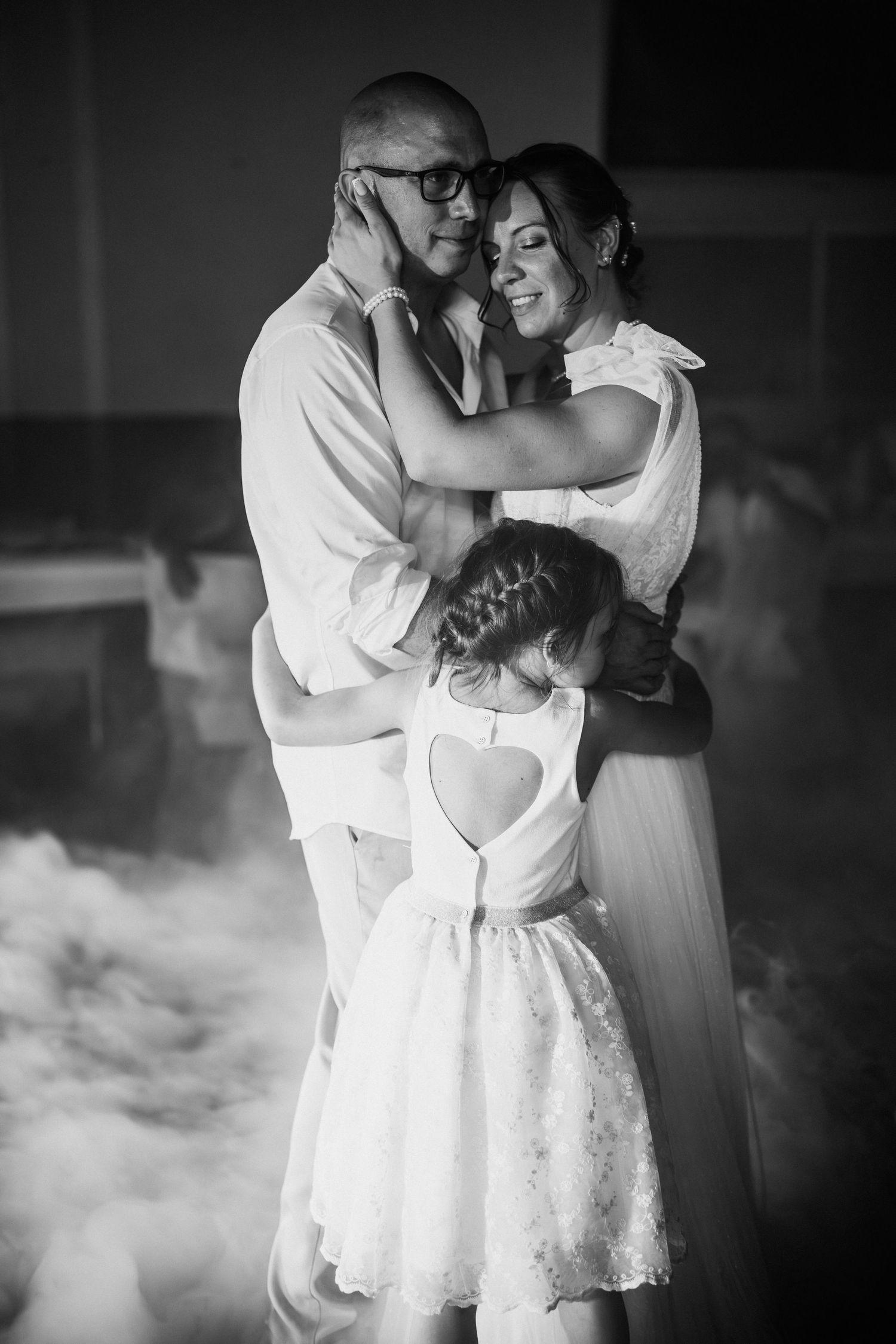 Black and white photo of a couple embracing on a dance floor with a young girl joining their hug at a wedding.
