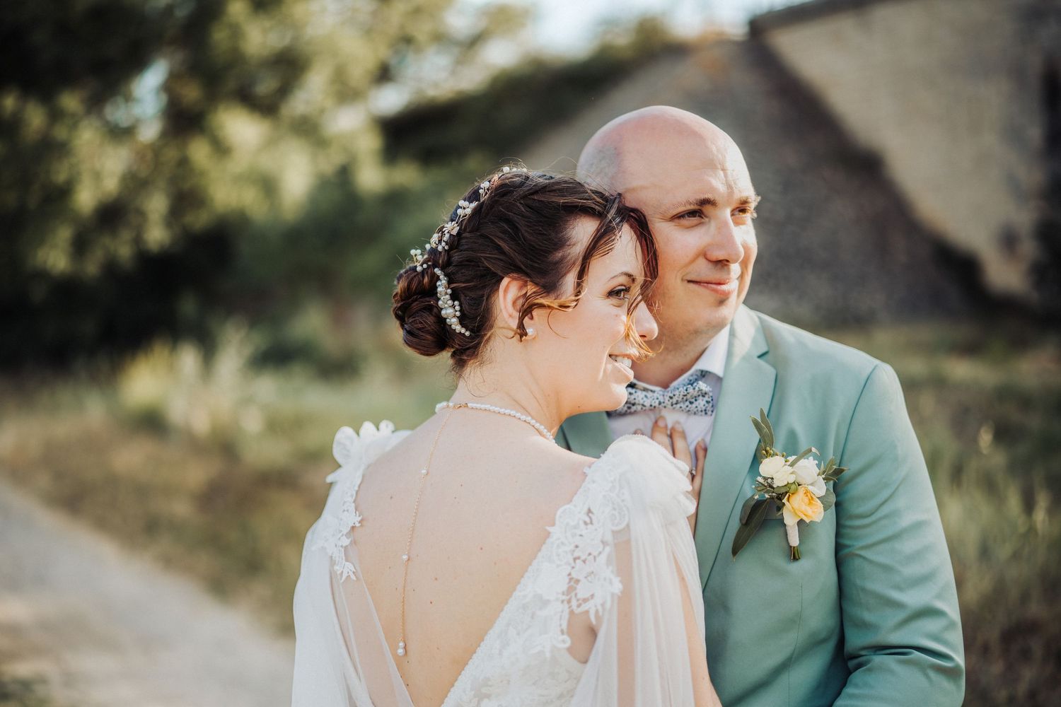Bride and groom posing outdoors at golden hour, bride in lace backless gown, groom in mint suit with floral boutonniere.
