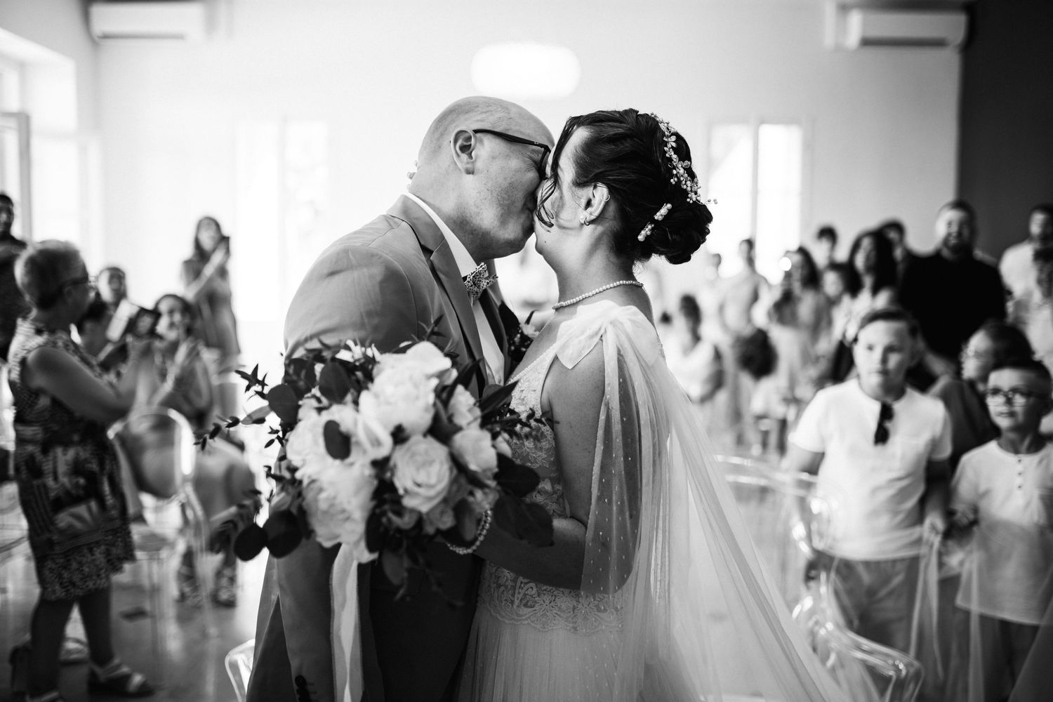 Black and white photo of a couple sharing their first kiss at a wedding ceremony with guests applauding in the background.