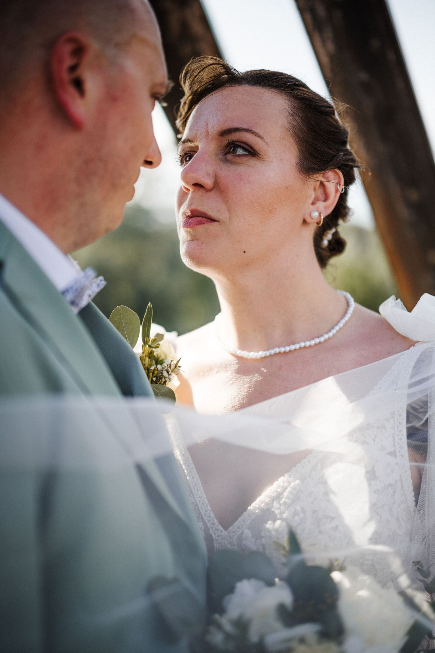 Bride in white lace dress and pearl necklace gazes lovingly at groom in sage suit during outdoor wedding ceremony.