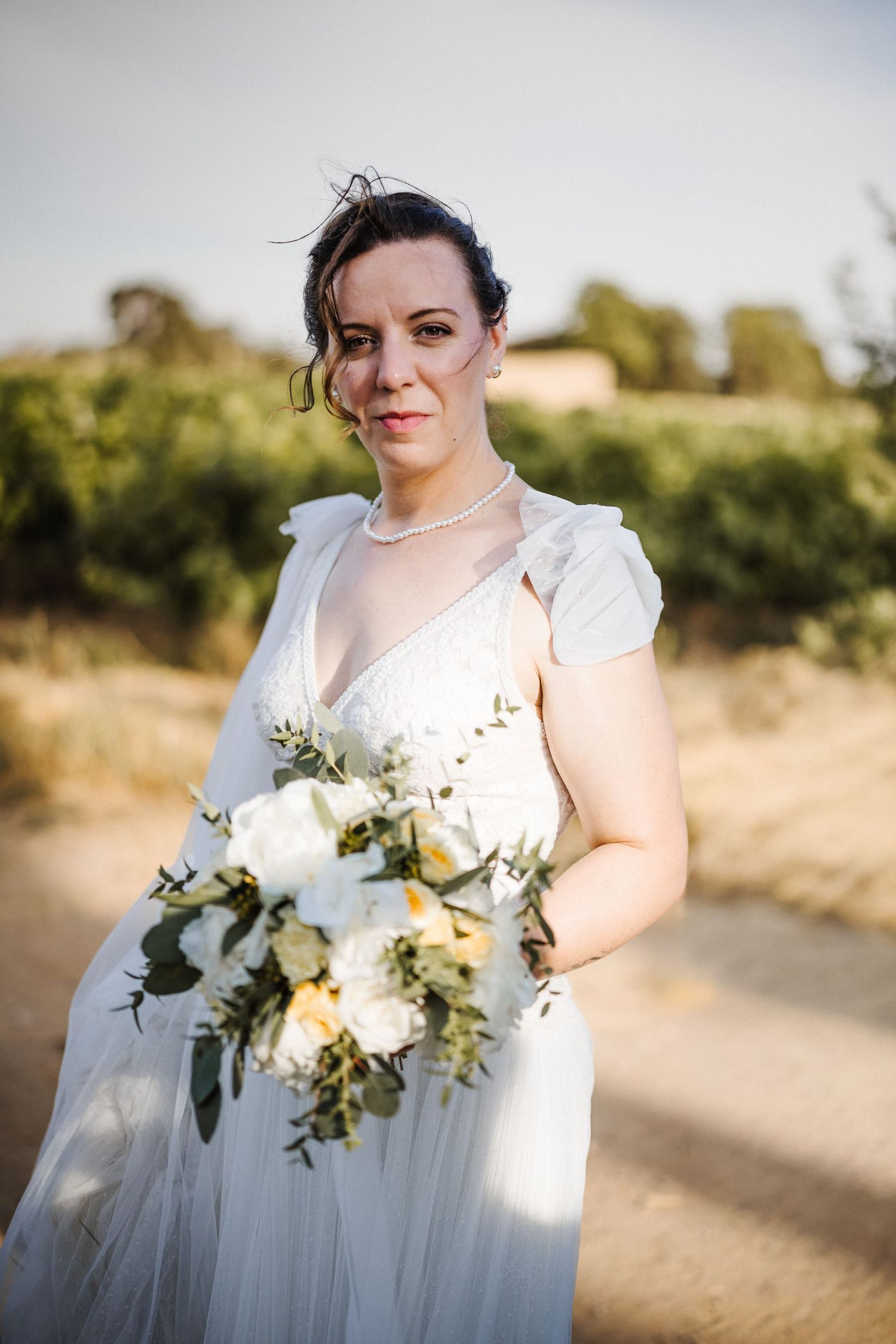 Bride in white gown holding white floral bouquet with greenery, standing on dirt path with vineyard background.