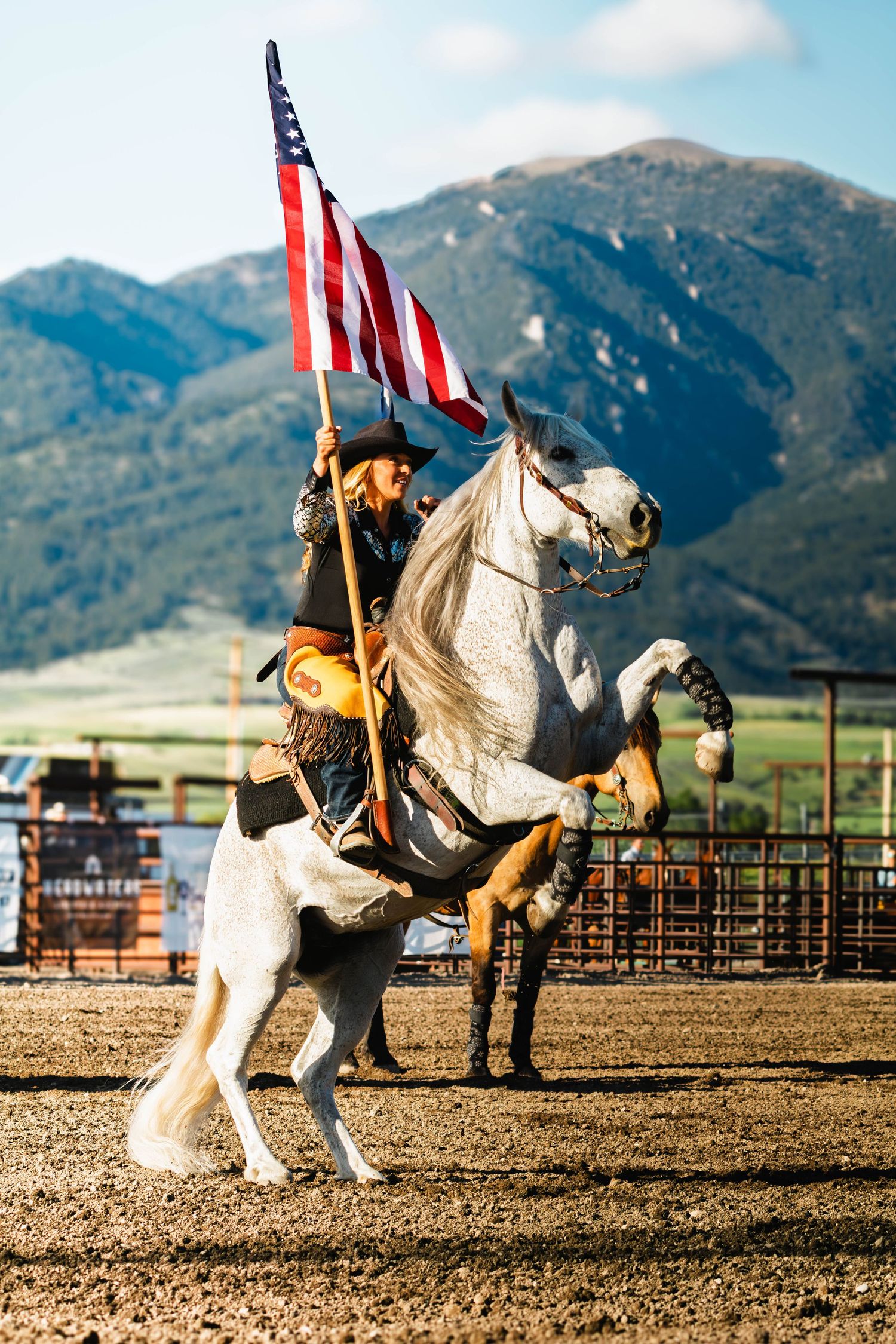Cowgirl on rearing white horse holding American flag at rodeo with mountain backdrop.