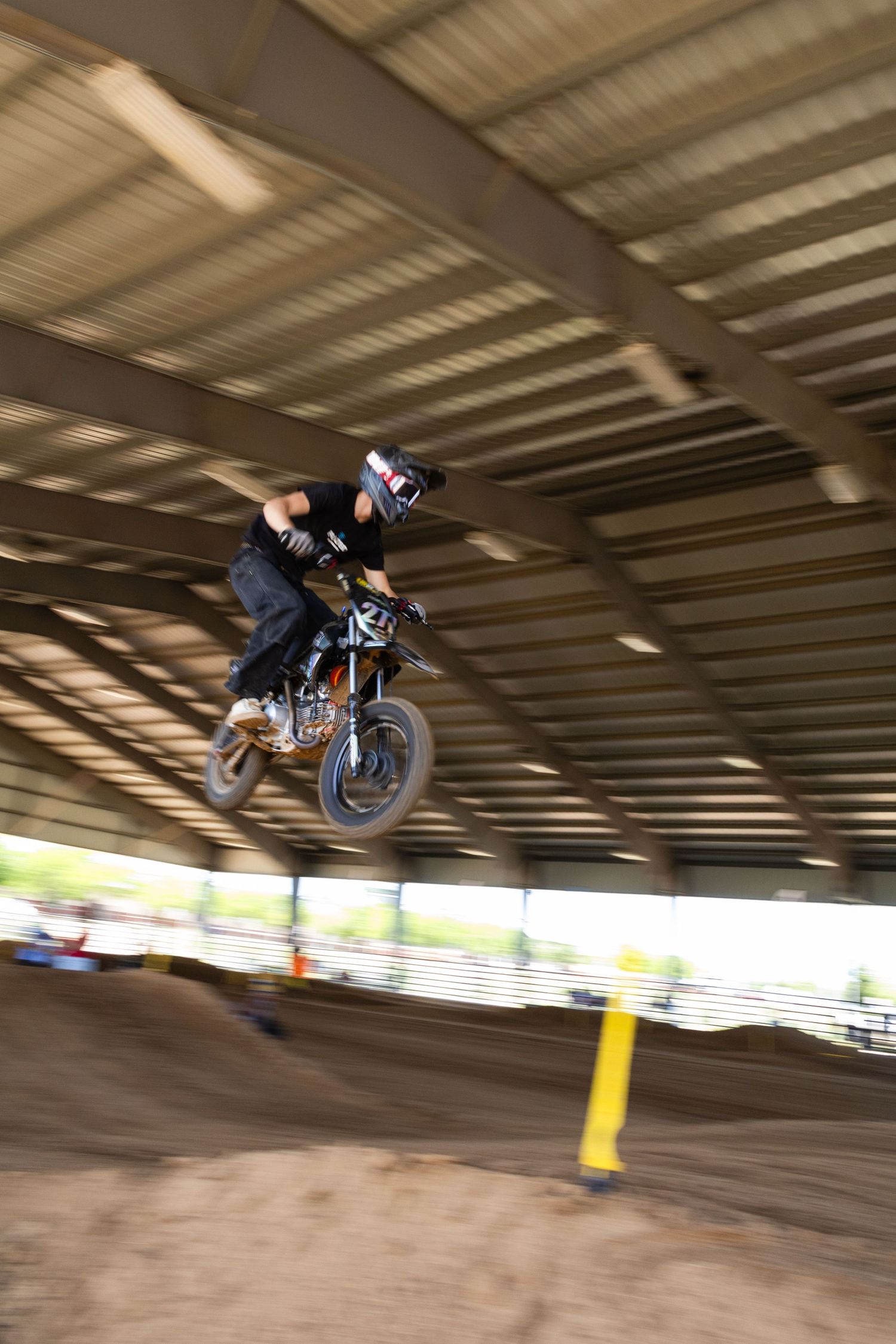 Motocross rider performing an aerial jump on a mini bike inside a covered dirt track arena.