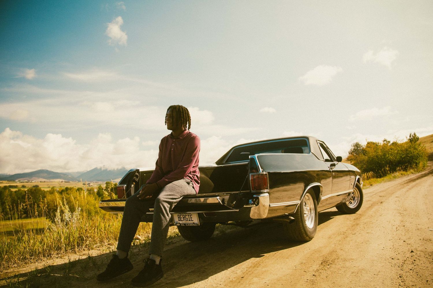 Person sitting on the trunk of a classic black car on a dirt road with mountain scenery behind.